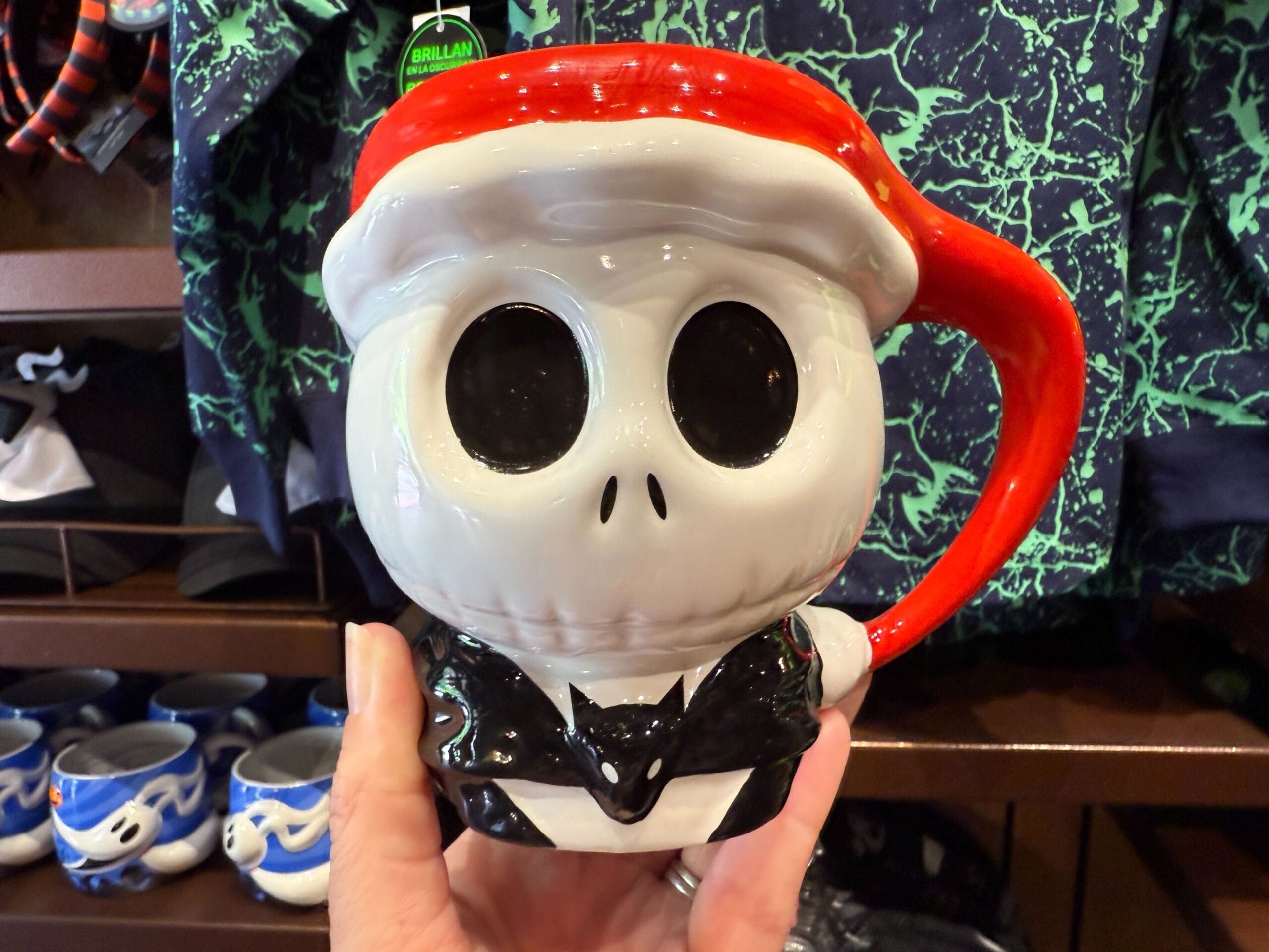 A hand holds a Jack Skellington Santa mug, classic Nightmare Before Christmas merchandise, in front of a shelf with other mugs.