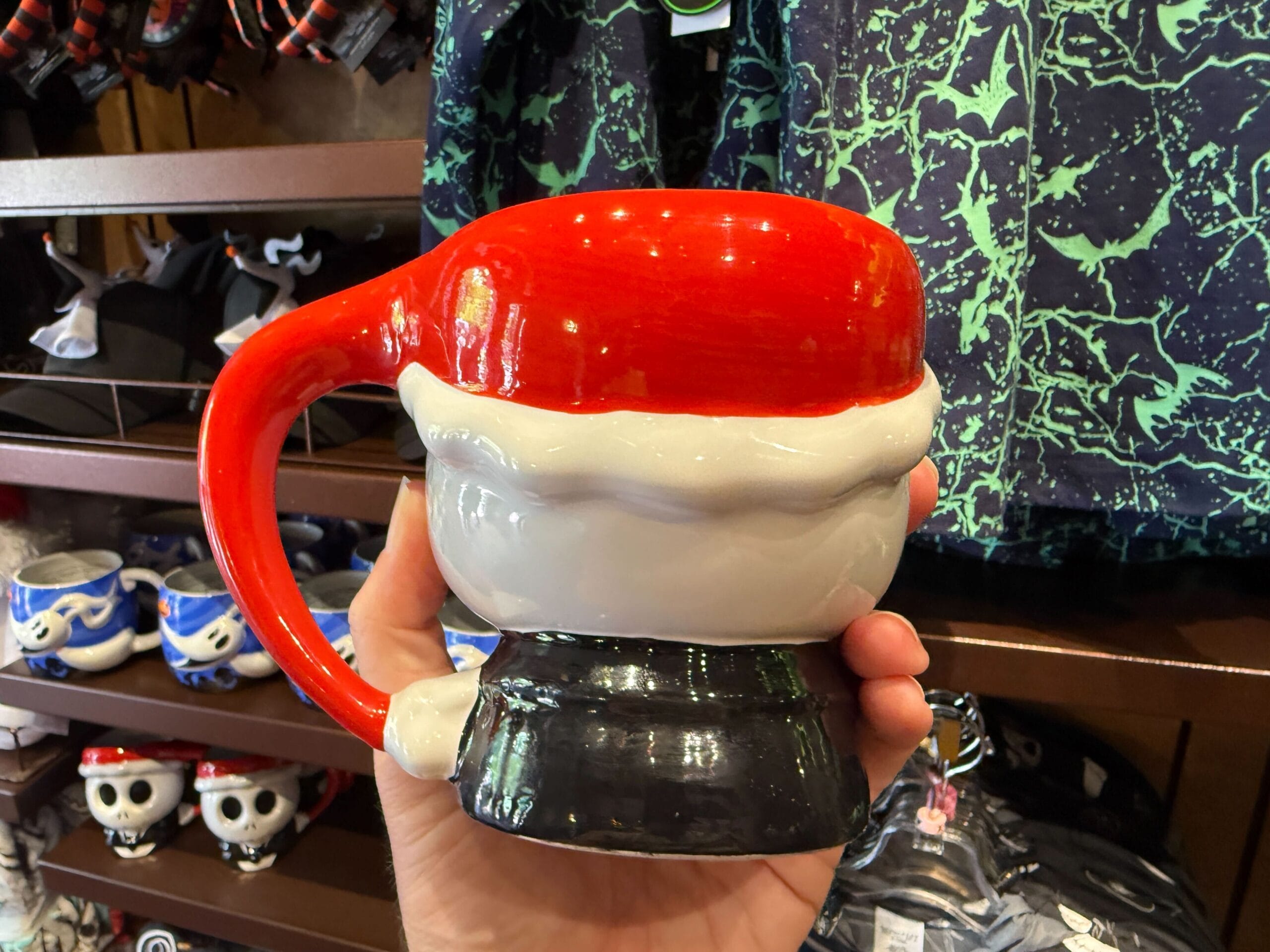 A hand holds a Santa hat mug amid merchandise, including The Nightmare Before Christmas novelty mugs on store shelves.