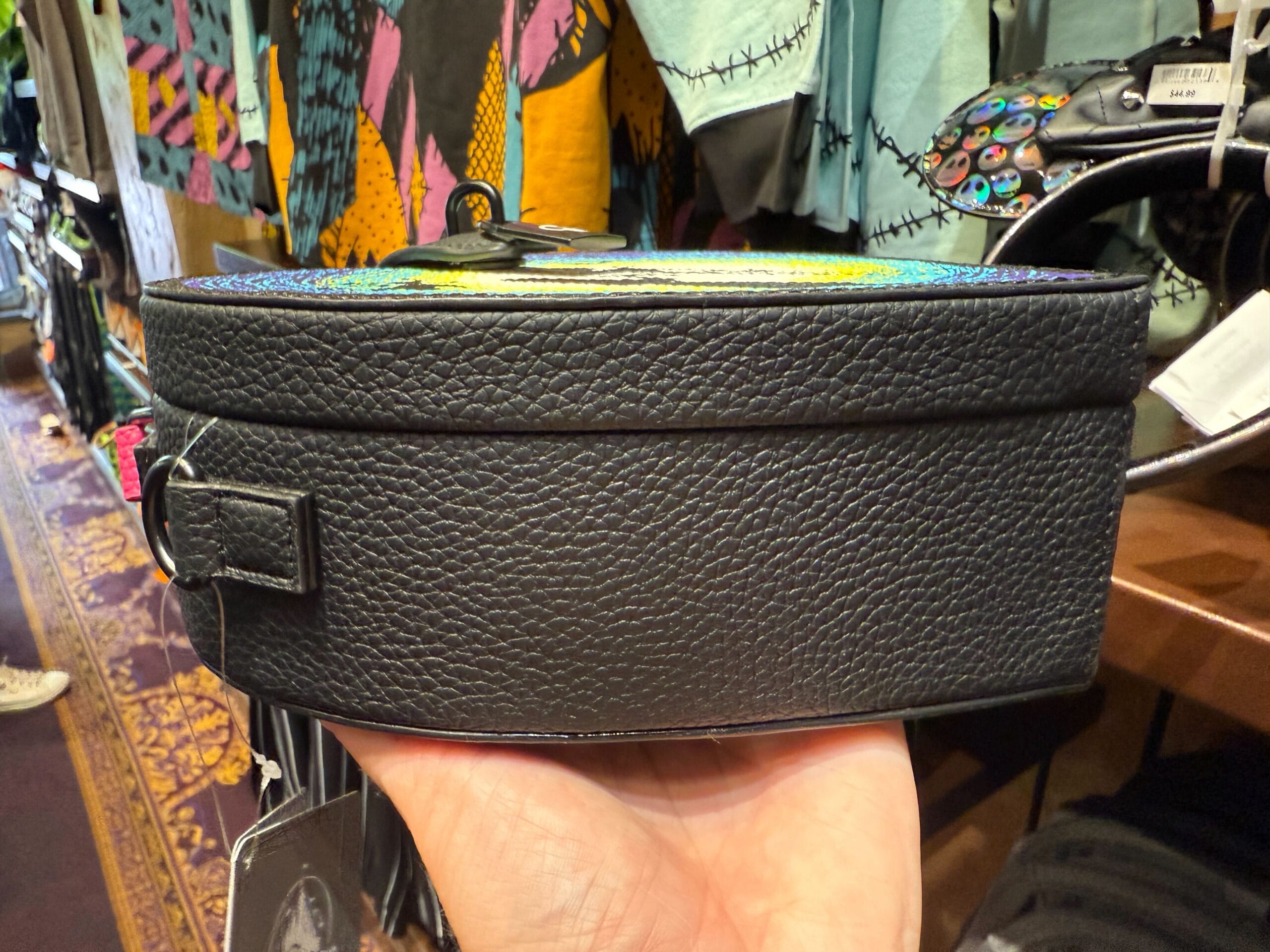 A hand holds a black, textured, oval-shaped The Nightmare Before Christmas merchandise bag in a store with colorful clothing.