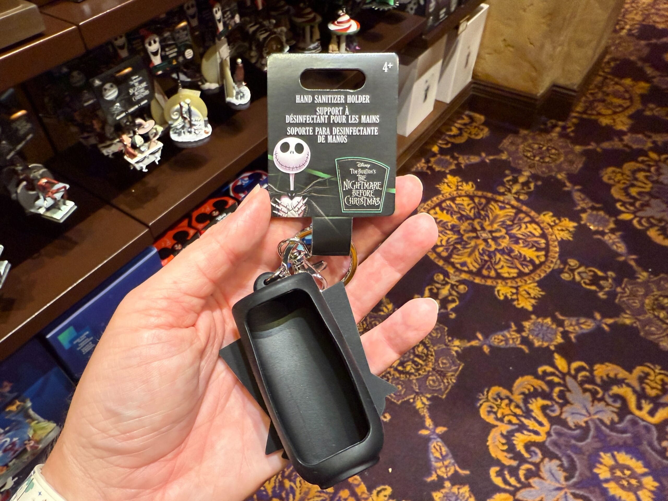A hand holds new Nightmare Before Christmas merchandise—a hand sanitizer holder—in a store with patterned carpet.