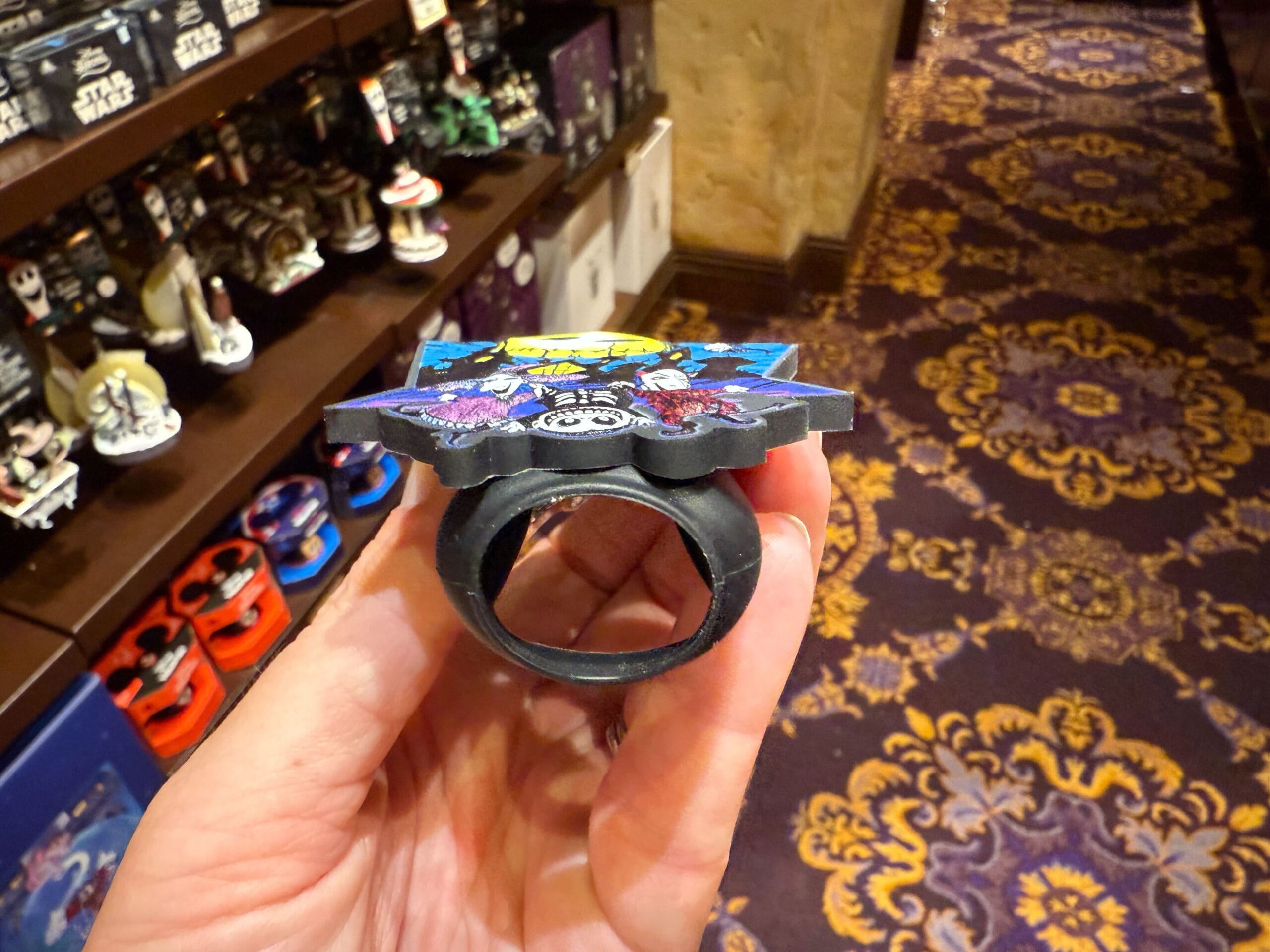 A hand holds a decorative ring in a store with Nightmare Before Christmas merchandise and patterned carpet in the background.
