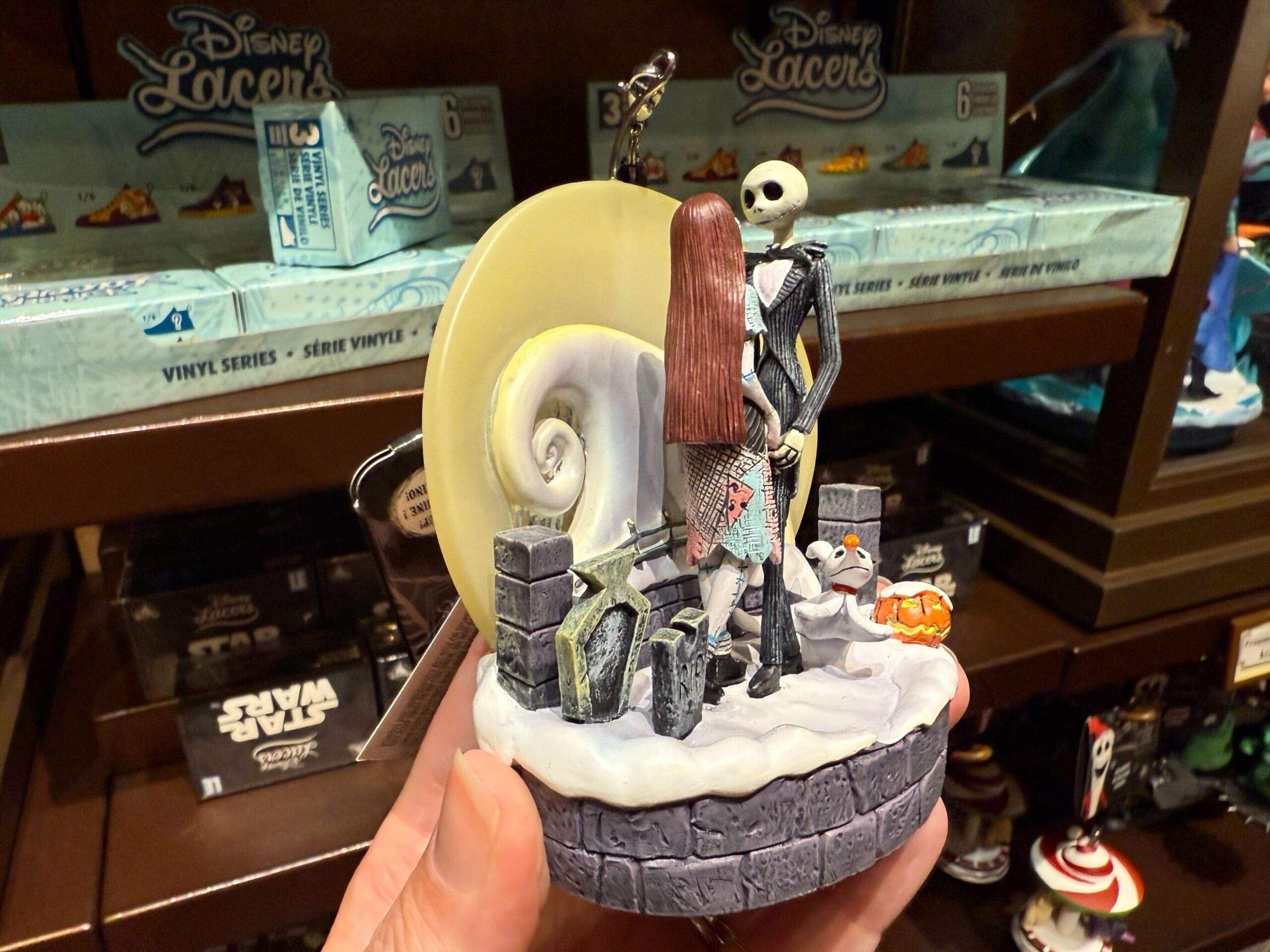 Hand holding The Nightmare Before Christmas merchandise ornament with Jack, Sally, Zero, and a moon background on a store shelf.