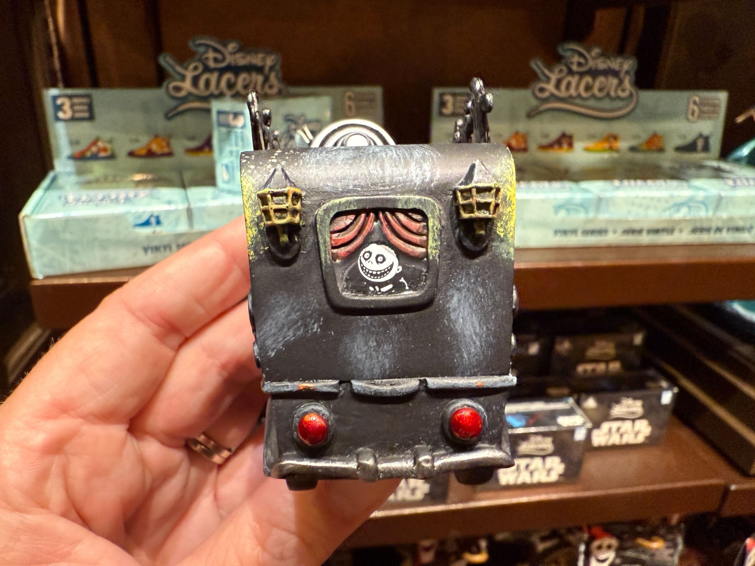 A hand holds a toy van with a Jack Skellington sticker, with Nightmare Before Christmas merchandise boxes in the background.