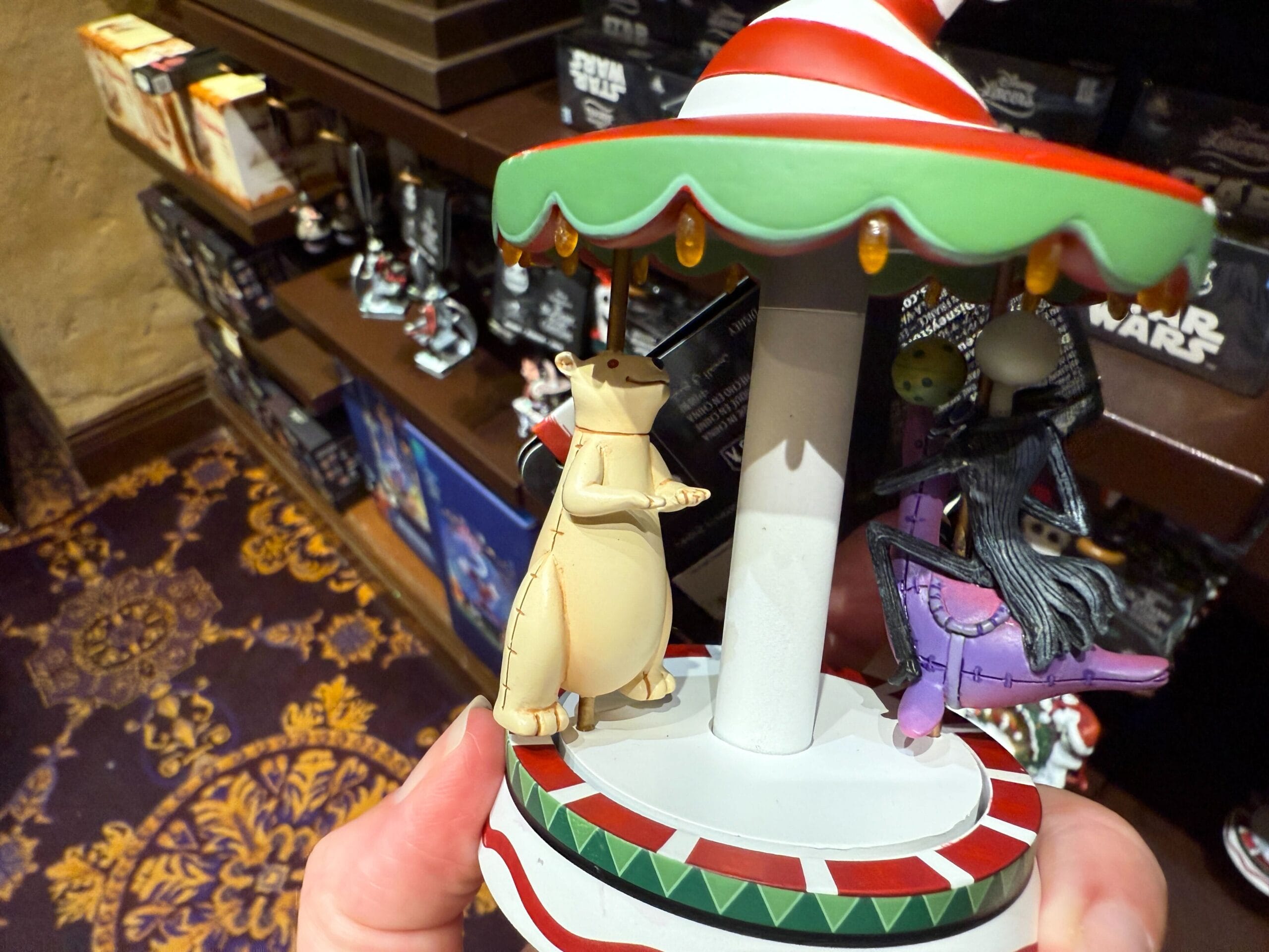 Hand holding The Nightmare Before Christmas carousel ornament, with animated characters, on merchandise-filled store shelves.