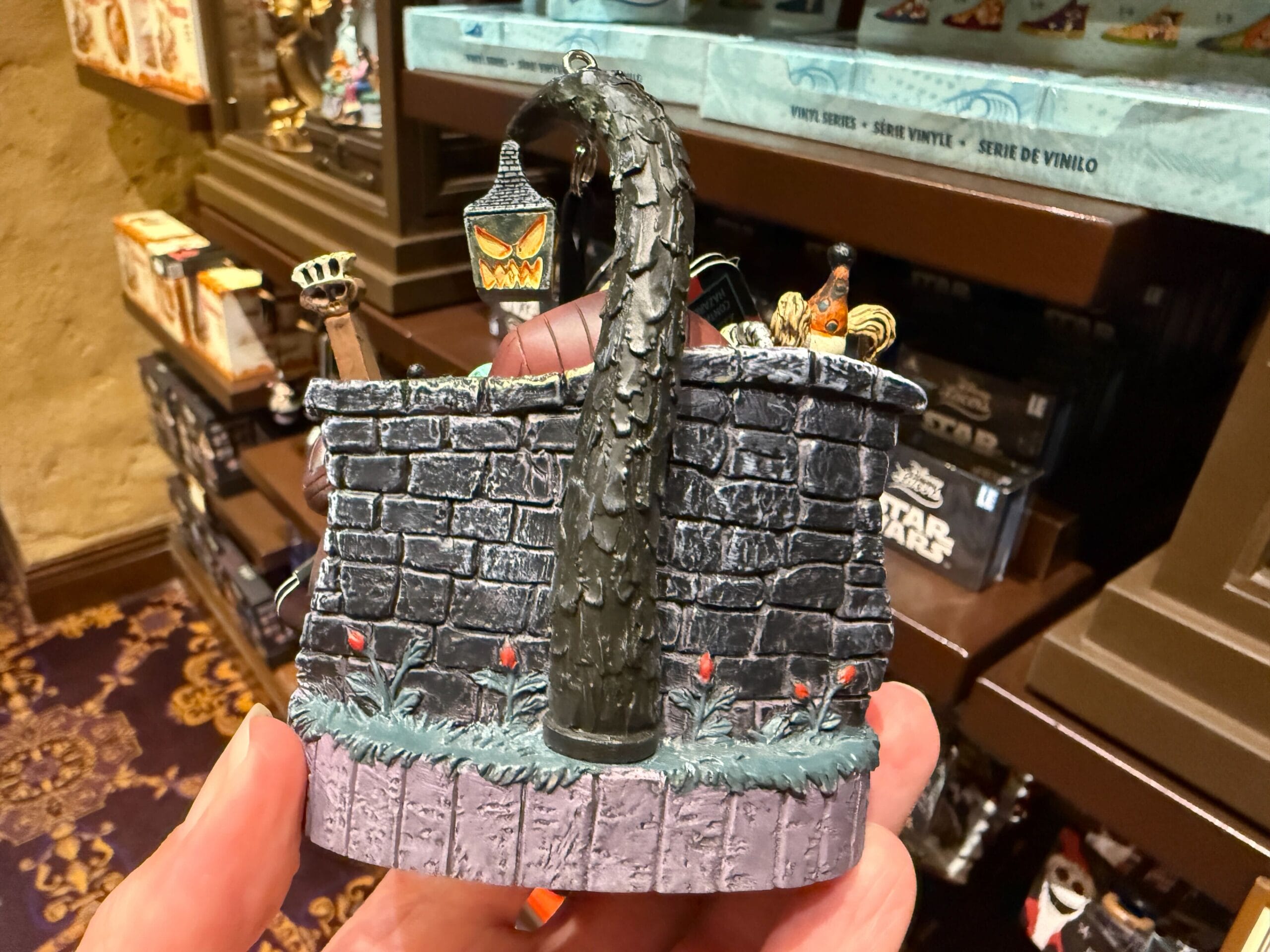 A hand holds The Nightmare Before Christmas merchandise figurine with a stone wall, lamp, and spooky tree in a store.