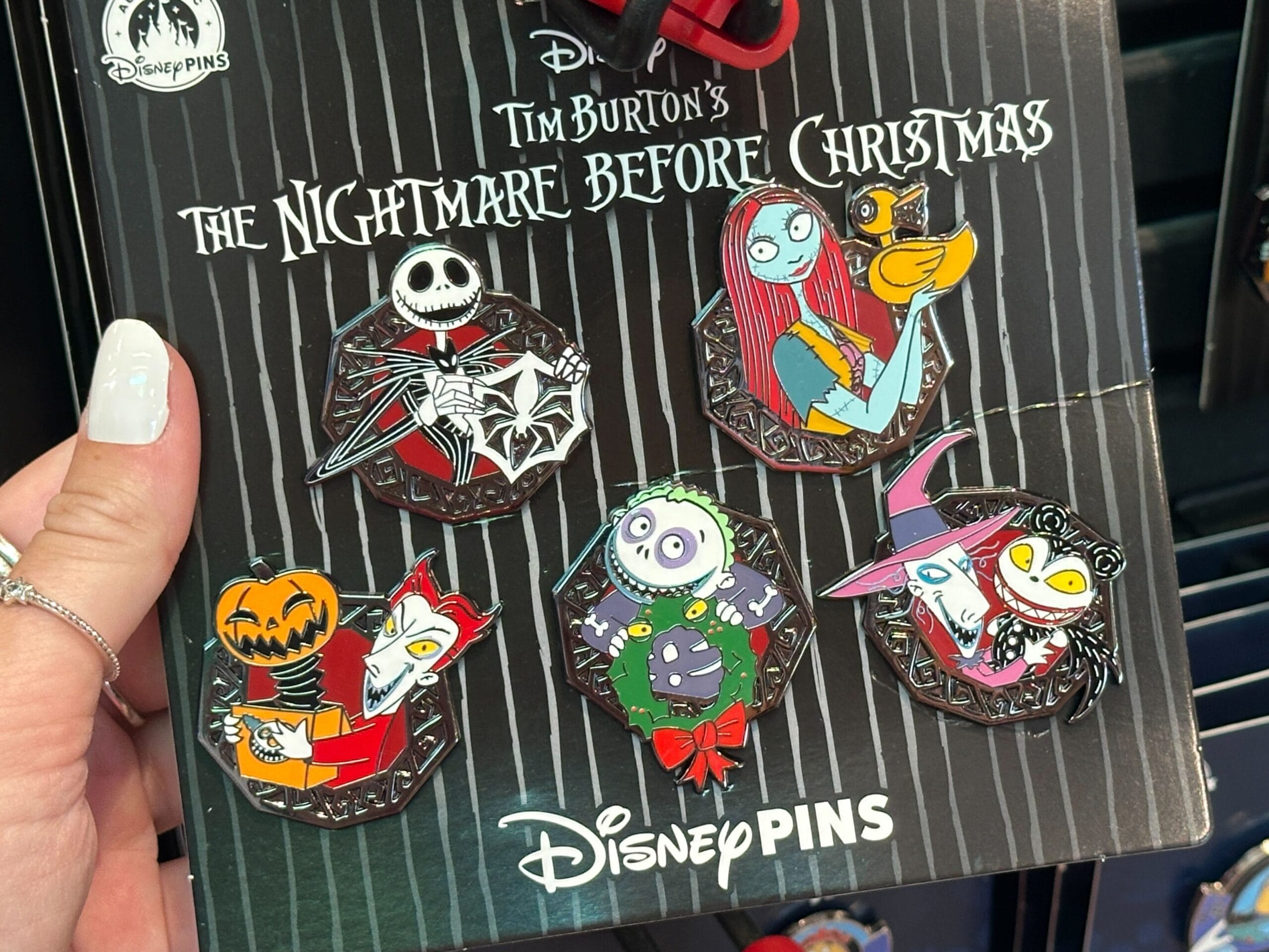 A hand holds six Nightmare Before Christmas merchandise pins featuring characters on a black Disney display card.