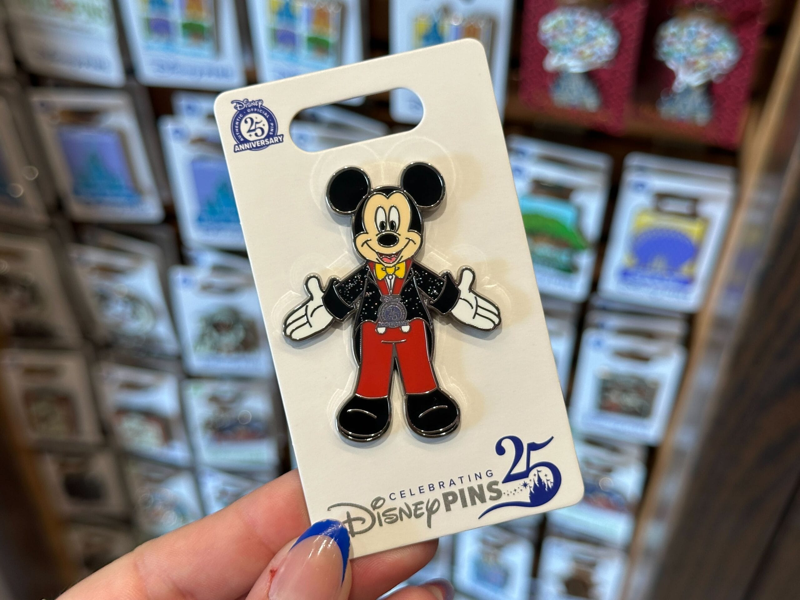 A hand holds a Disney pin of Mickey Mouse in red pants, celebrating the 25th anniversary of Disney Pins.