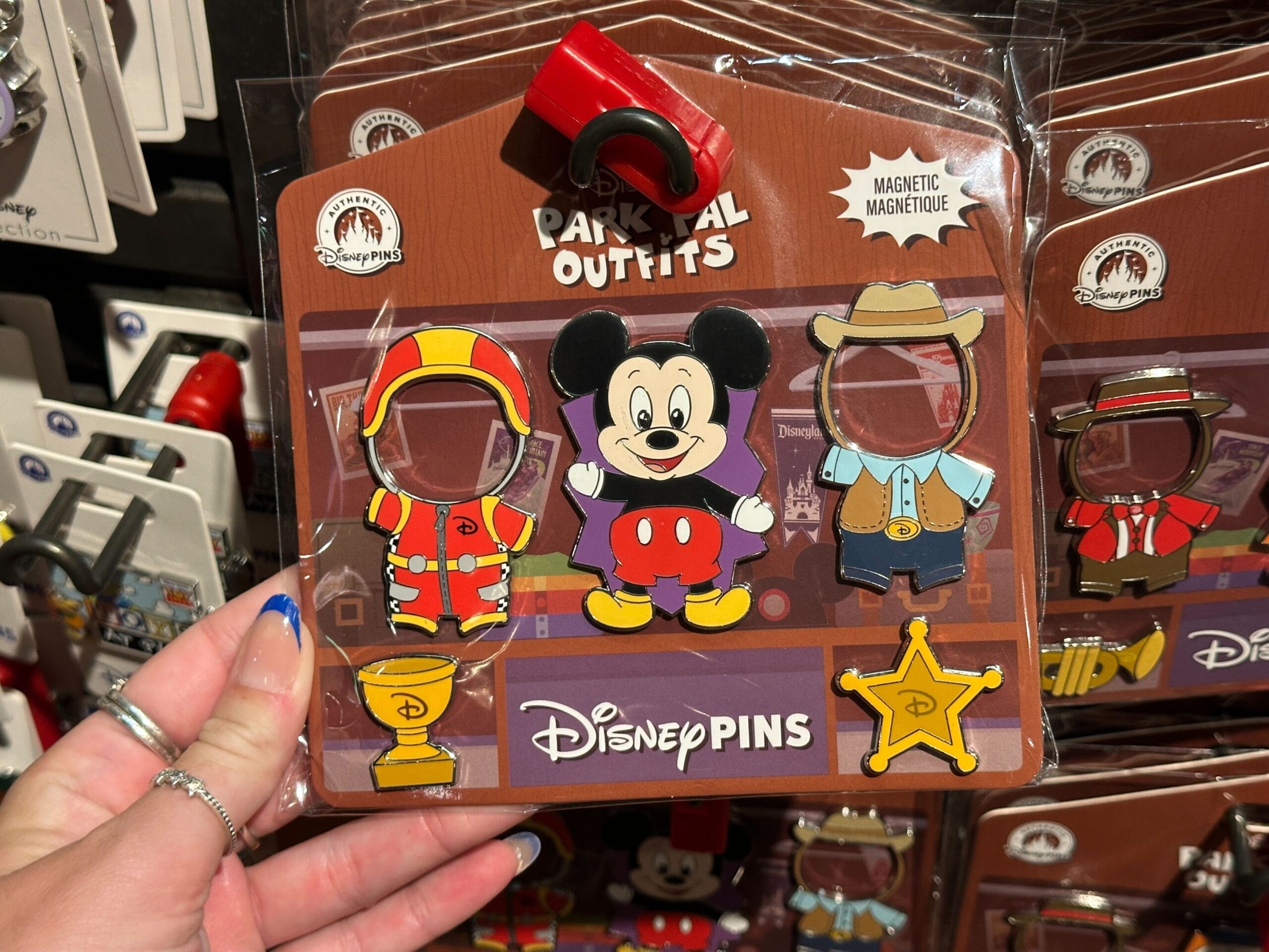 Hand holding Disney pins with Mickey Mouse and magnetic park ranger and cowboy outfits on display rack.