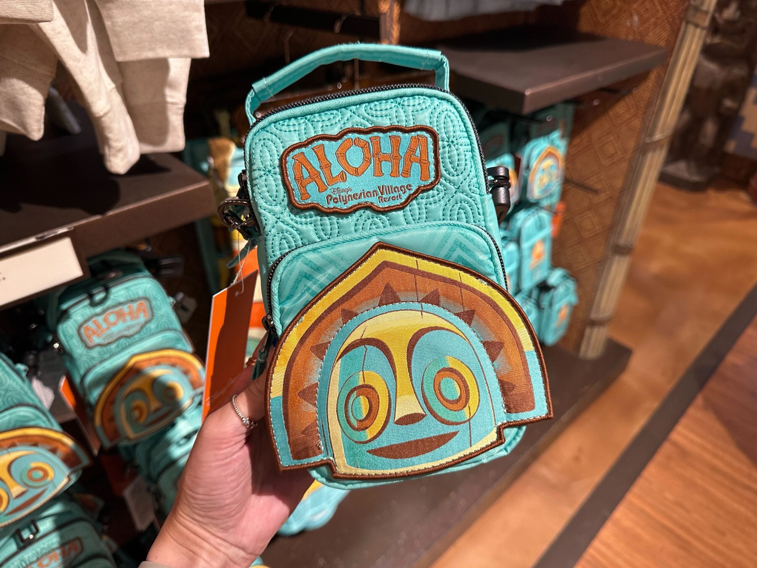 A hand holds a teal bag with "Aloha" and a tiki face design, displayed in a store with similar bags in the background.