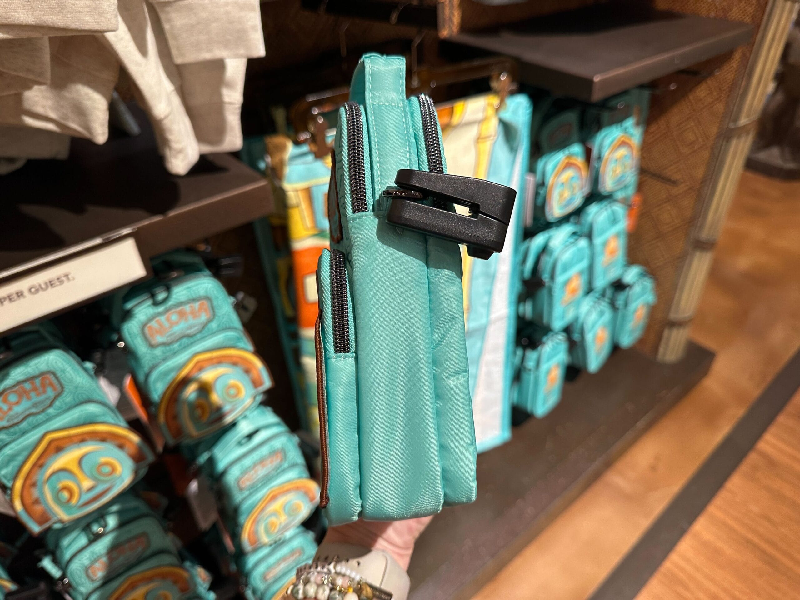 A hand holds a small teal bag with a zipper and a black clip, displayed in a store with similar bags in the background.