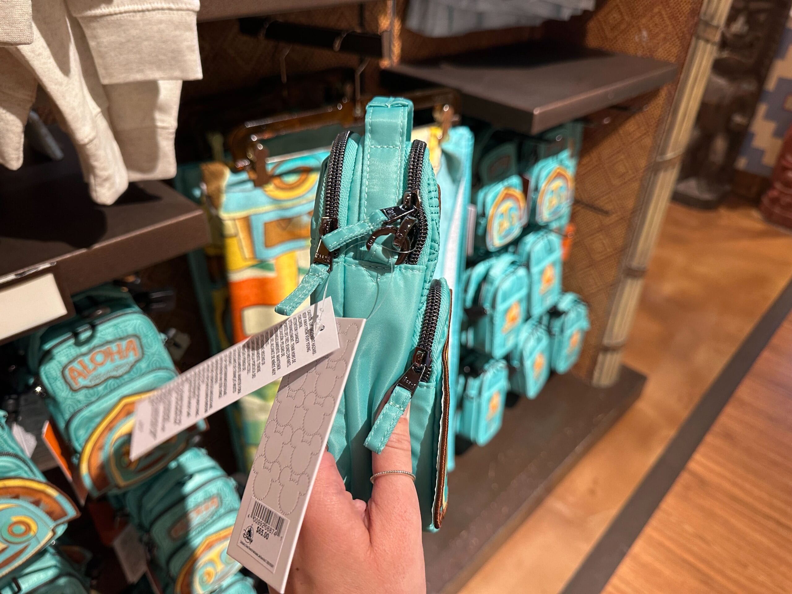 A hand holds a teal zippered pouch in a store, with similar bags displayed on shelves in the background.