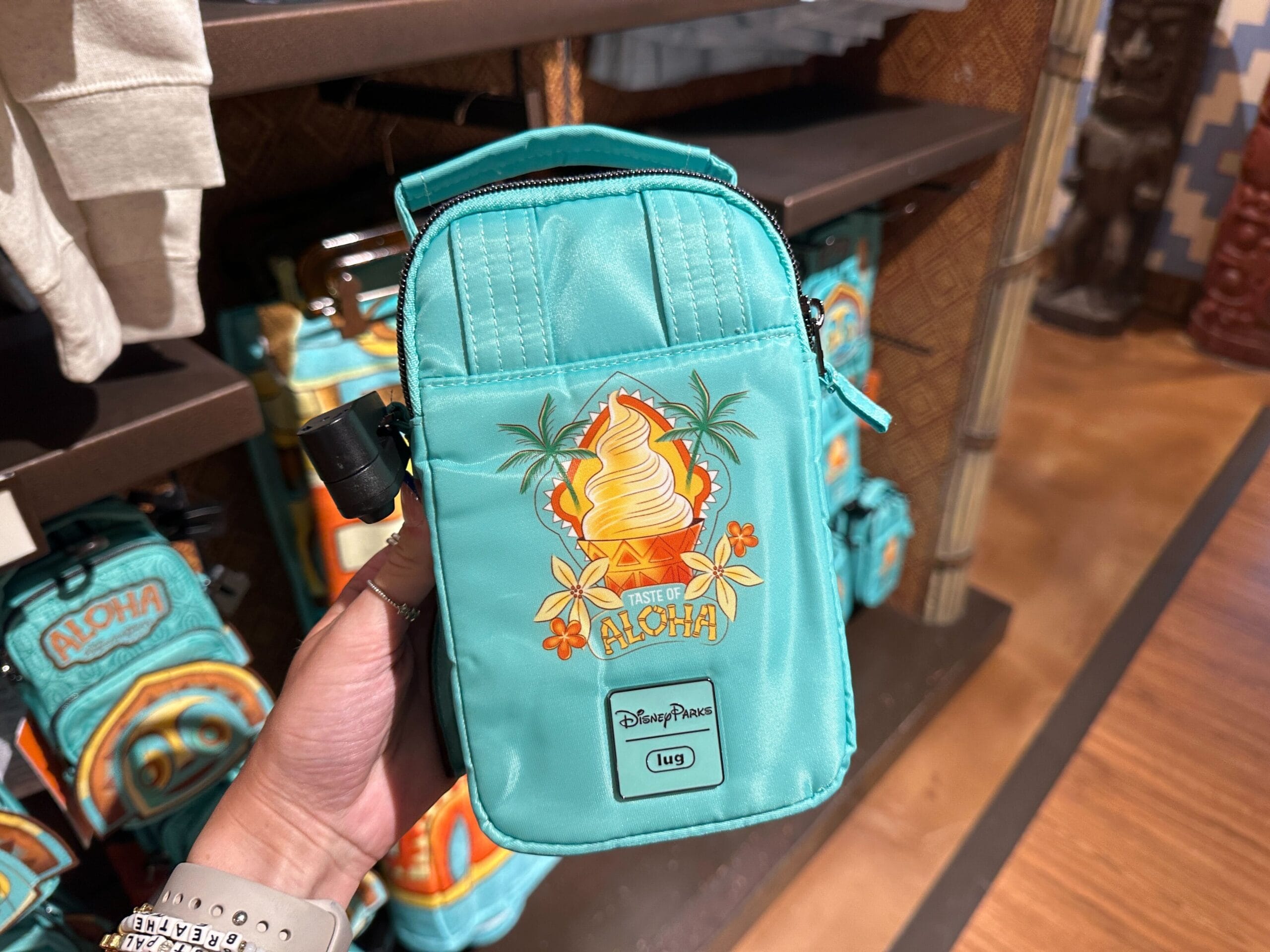 A teal Disney Parks pouch with a Dole Whip and "Taste of Aloha" design held in a hand in a store.