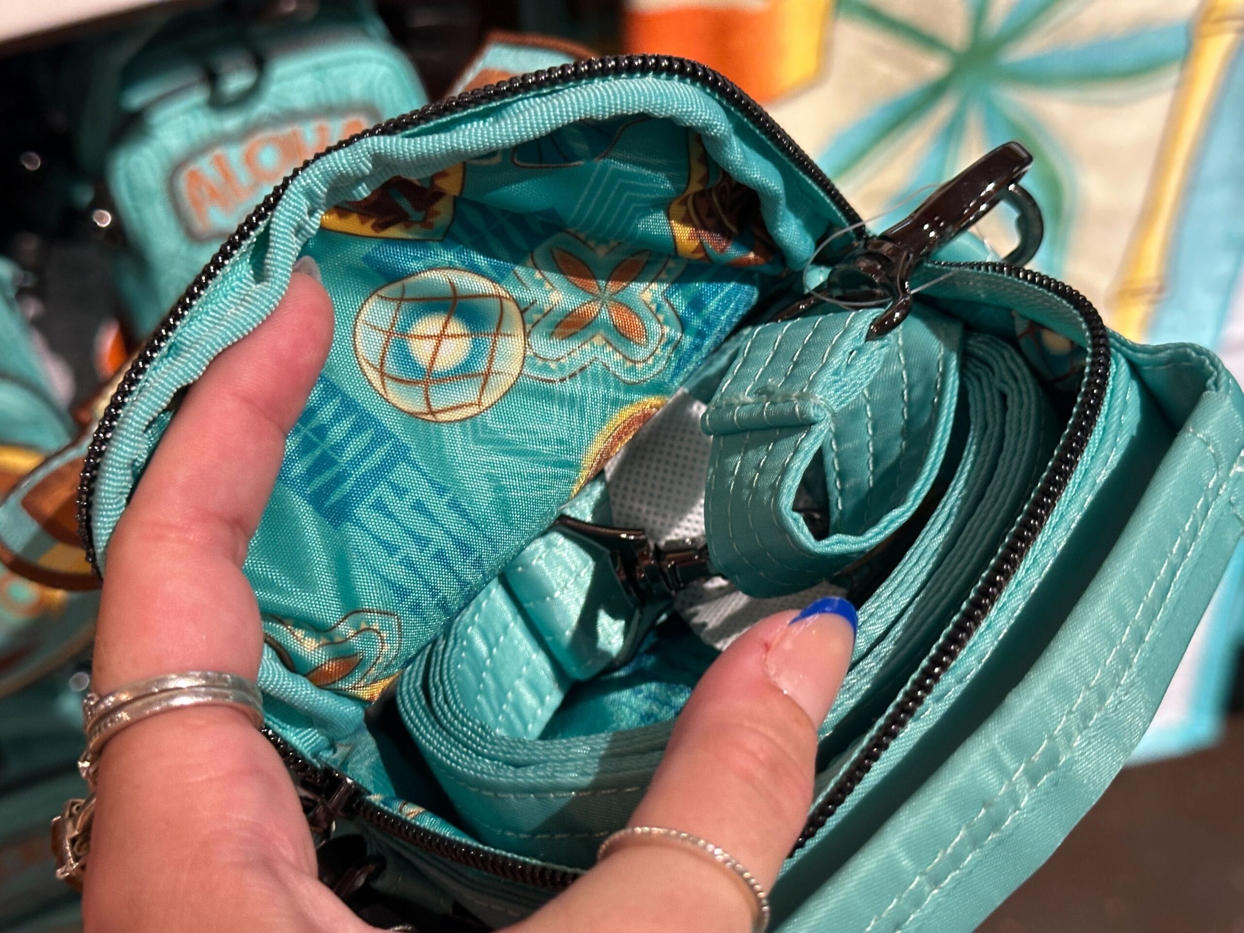 A hand with rings holds open a teal bag, showing a patterned fabric lining with tropical designs inside.
