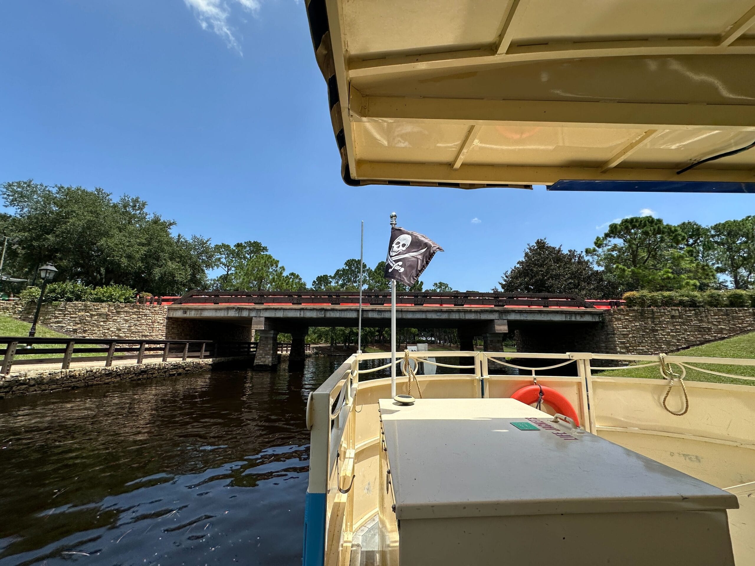 View from a boat with a pirate flag, cruising under a bridge near Disney’s Port Orleans Resort on a sunny day.