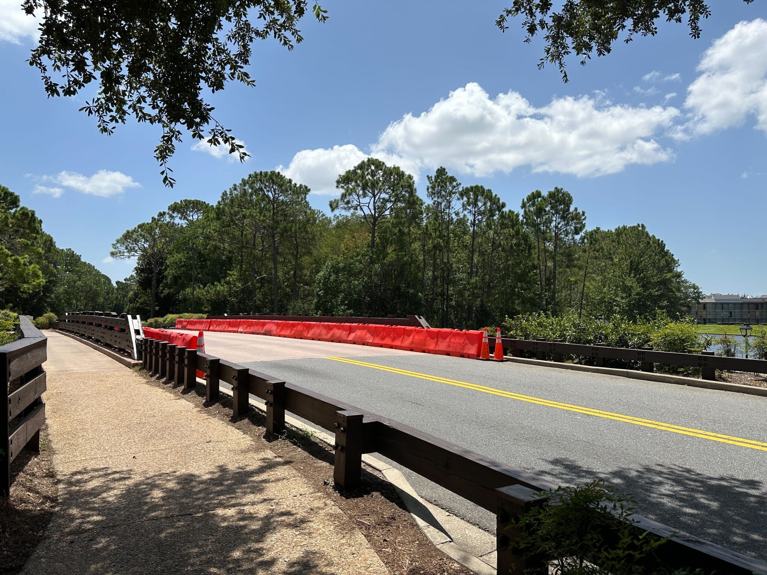 A road with orange barriers near Disney’s Port Orleans Resort runs beside a sidewalk and trees under a blue, cloud-scattered sky.