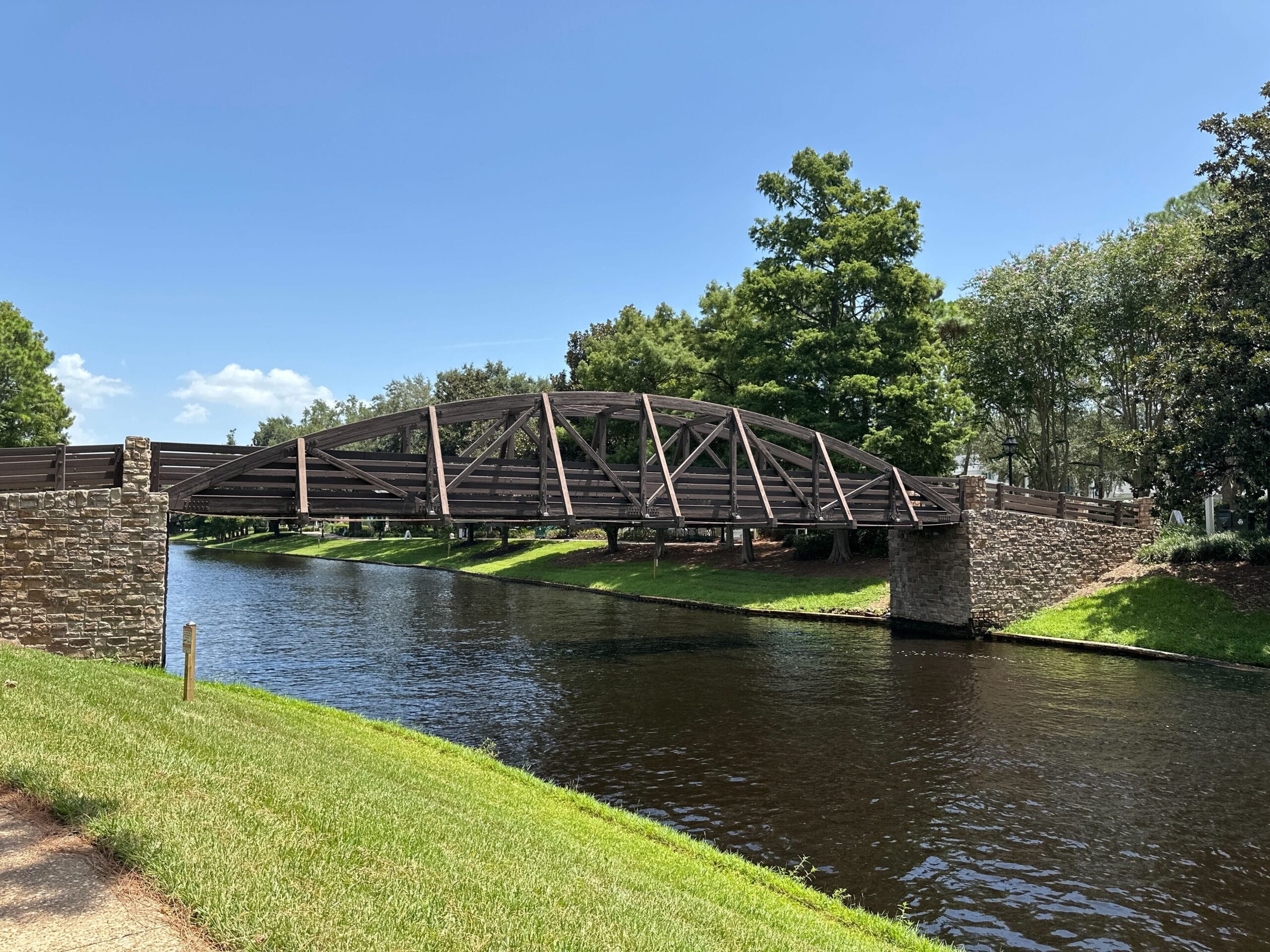 Brown metal and stone pedestrian bridge over a calm canal at Disney’s Port Orleans Resort under a clear blue sky.