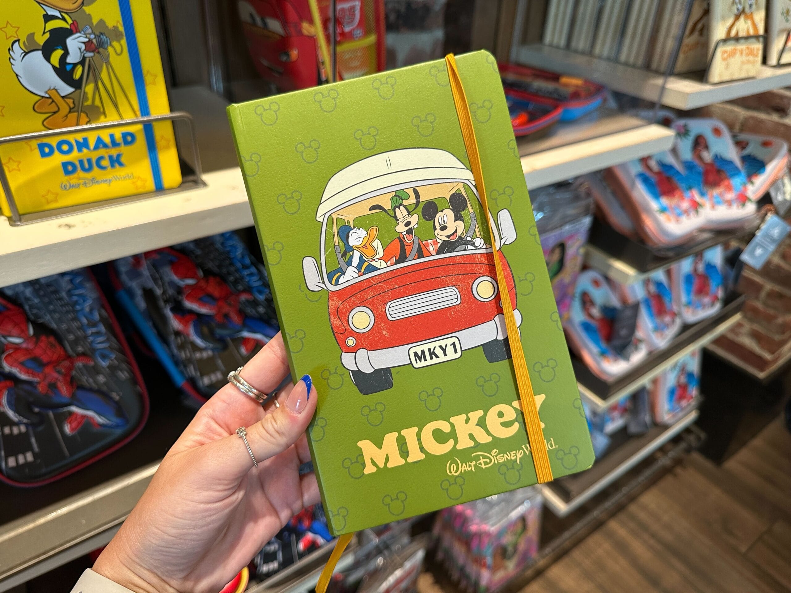 A hand holds a green Mickey Mouse notebook, part of Disney World merchandise, featuring characters in a red car on the cover.