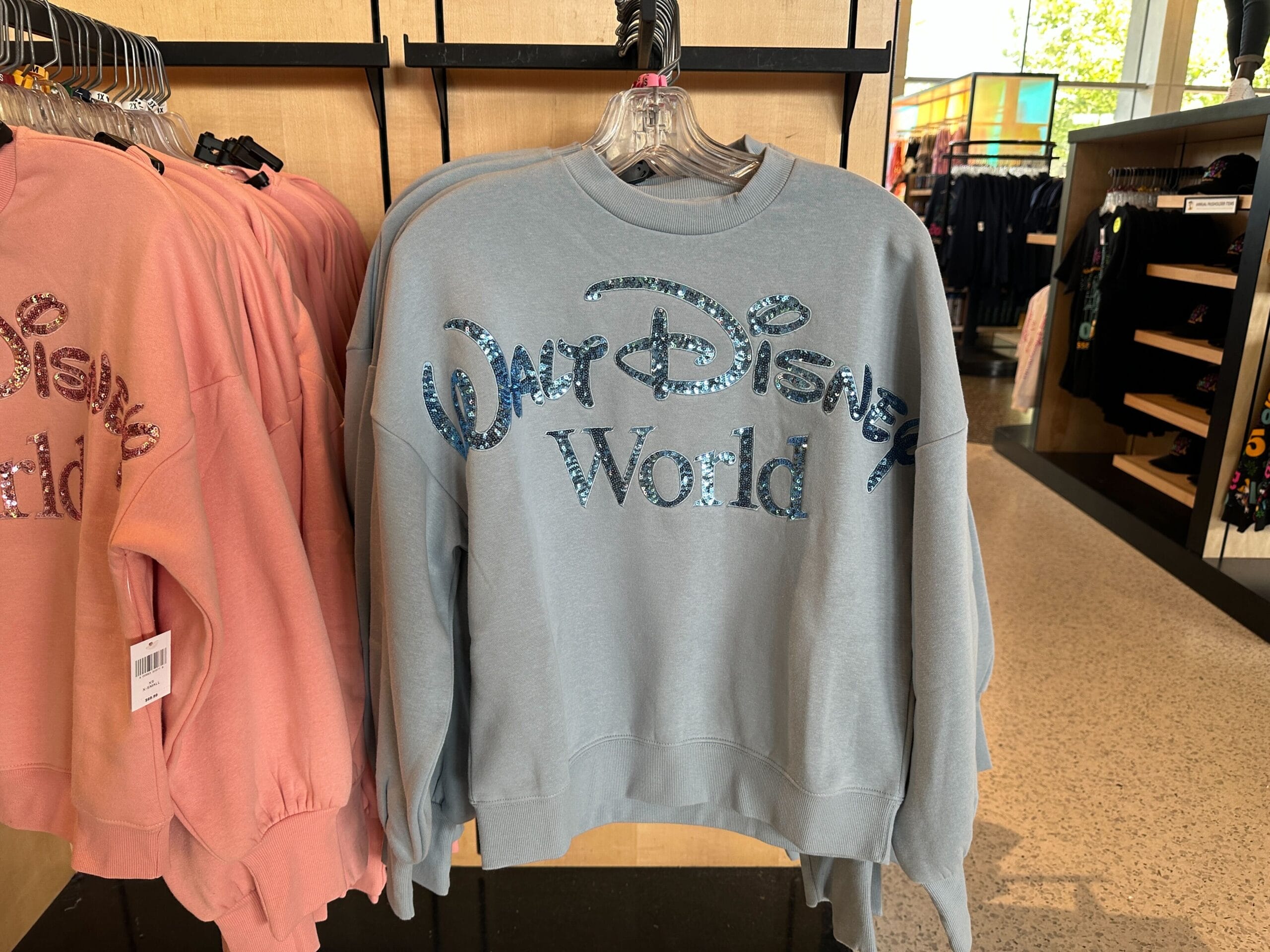 A light blue WDW sweatshirt with glittery lettering hangs on a store rack, perfect Disney merchandise for fans.