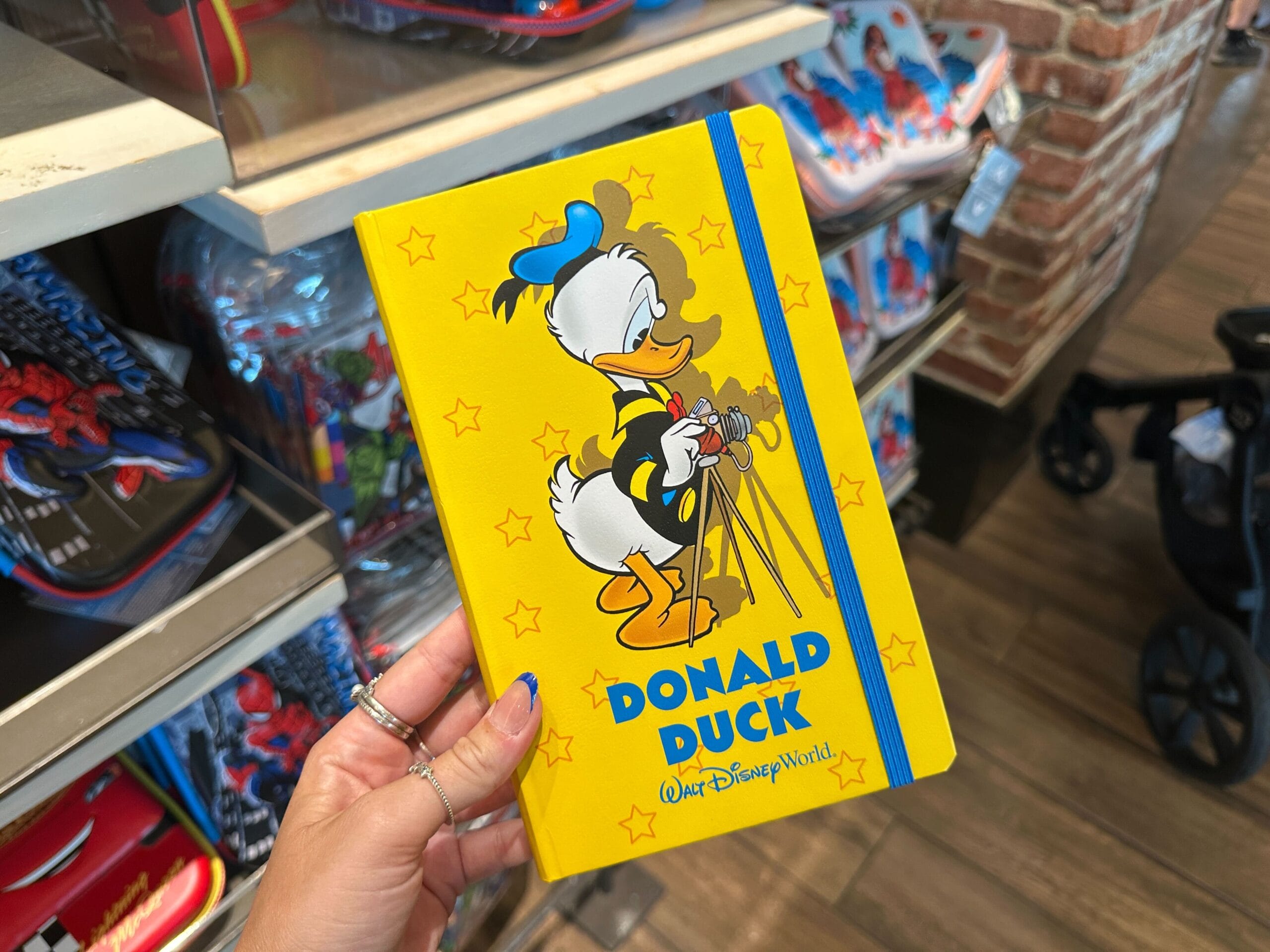 A hand holds a yellow Donald Duck notebook with stars, featured in a Walt Disney World merchandise display.