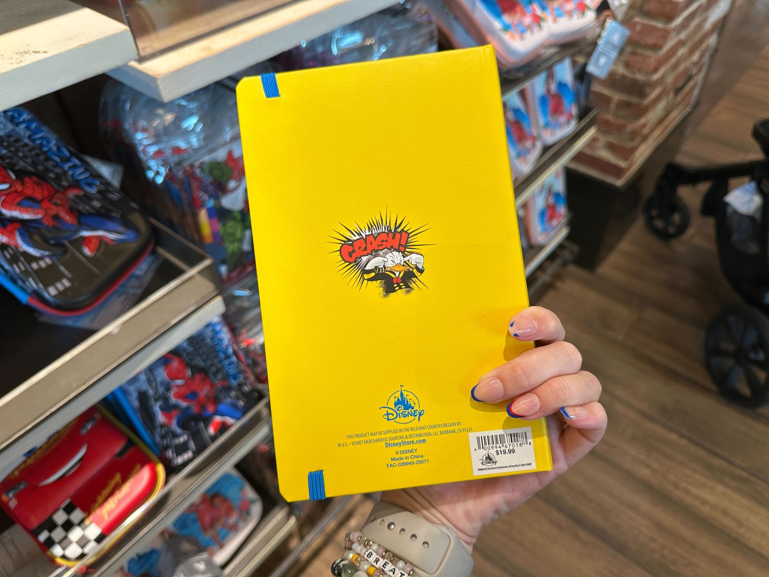 A hand holds a yellow Disney notebook with "CRASH!" graphic, displayed among WDW merch like lunchboxes on store shelves.
