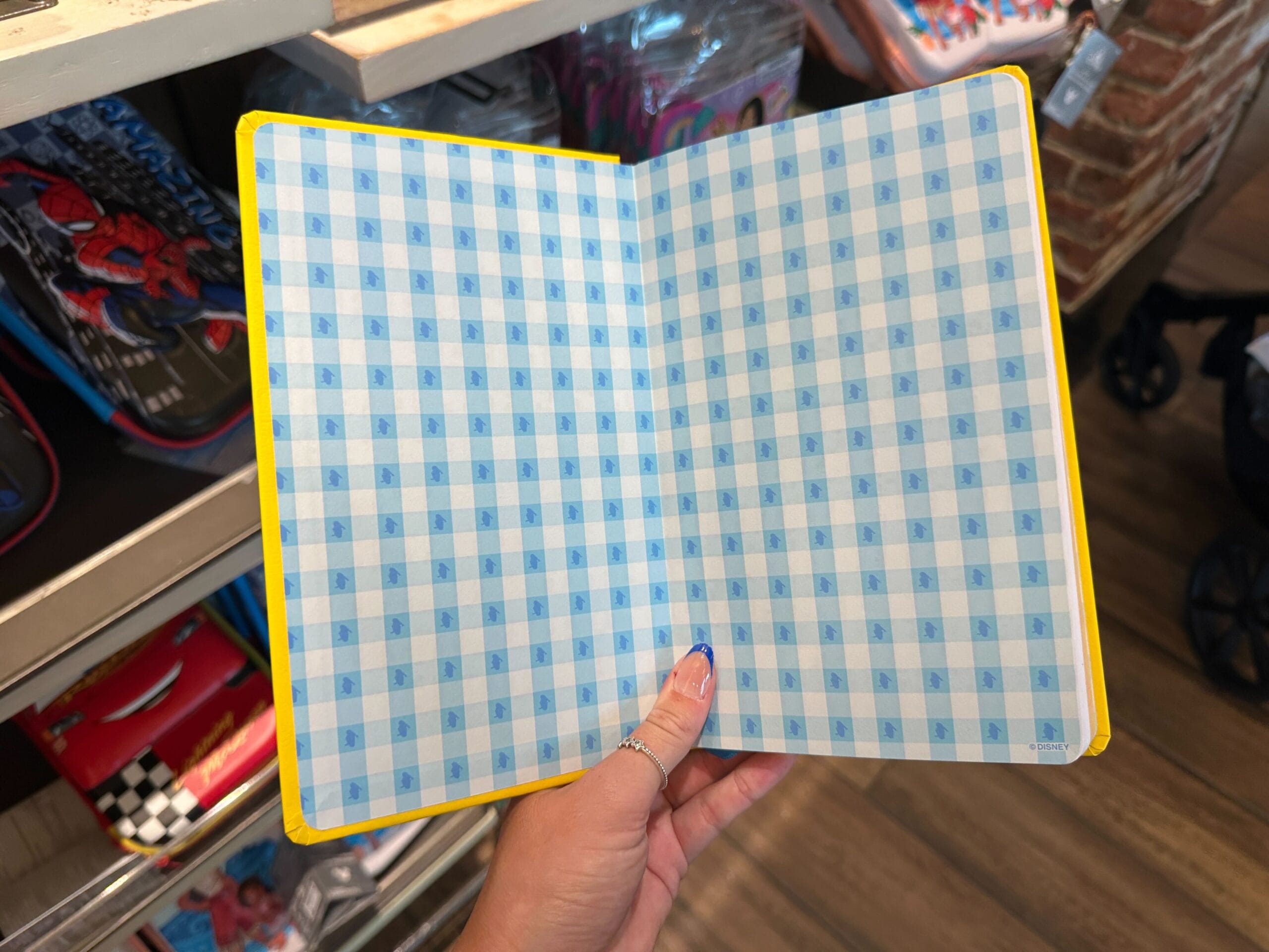 A hand holds open a Disney World Merchandise notebook with blue checkered pages and a yellow cover in a store.