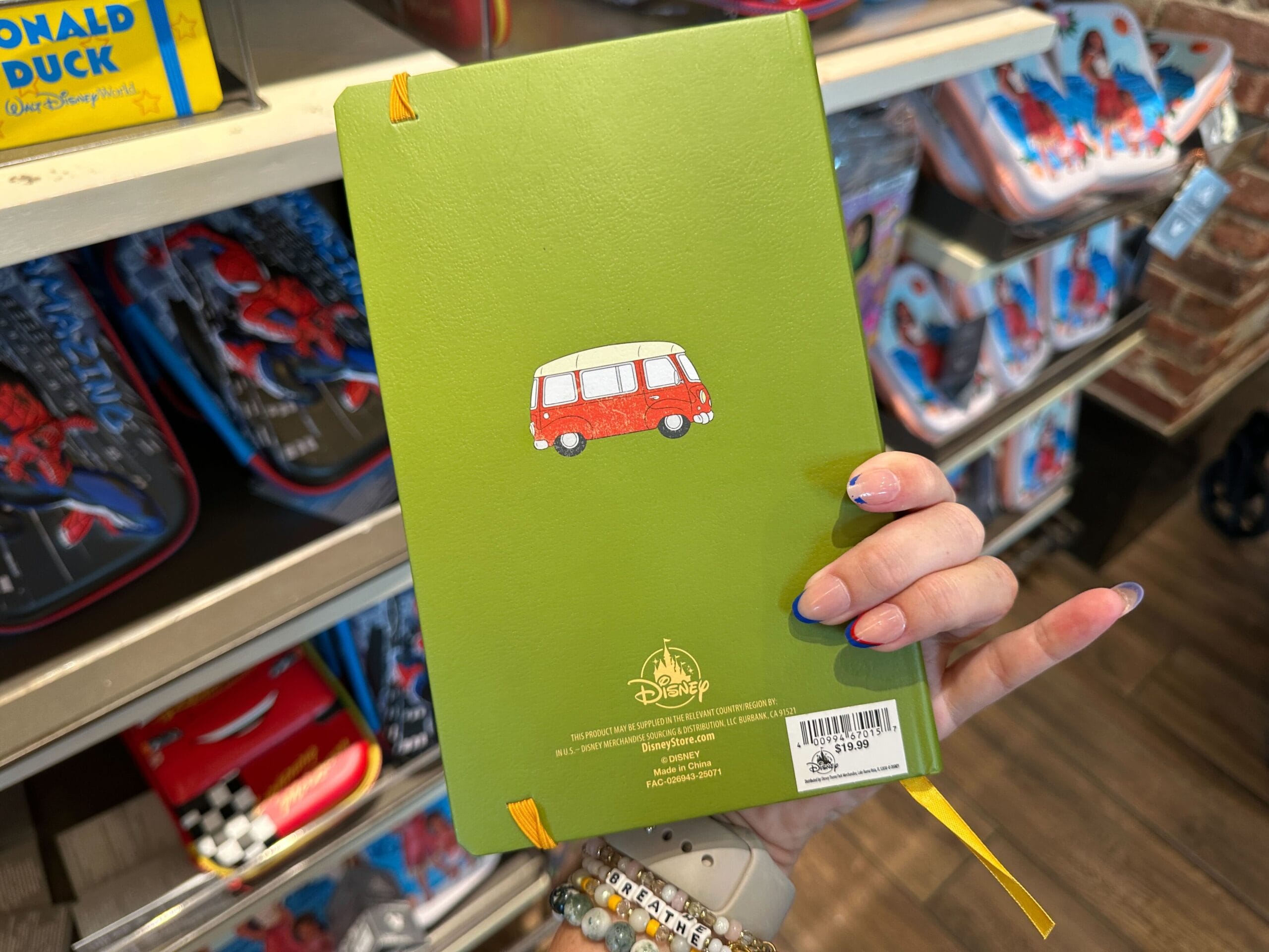 A hand holds a green Disney notebook with a van illustration—perfect as WDW merch or Disney souvenir—in a store.
