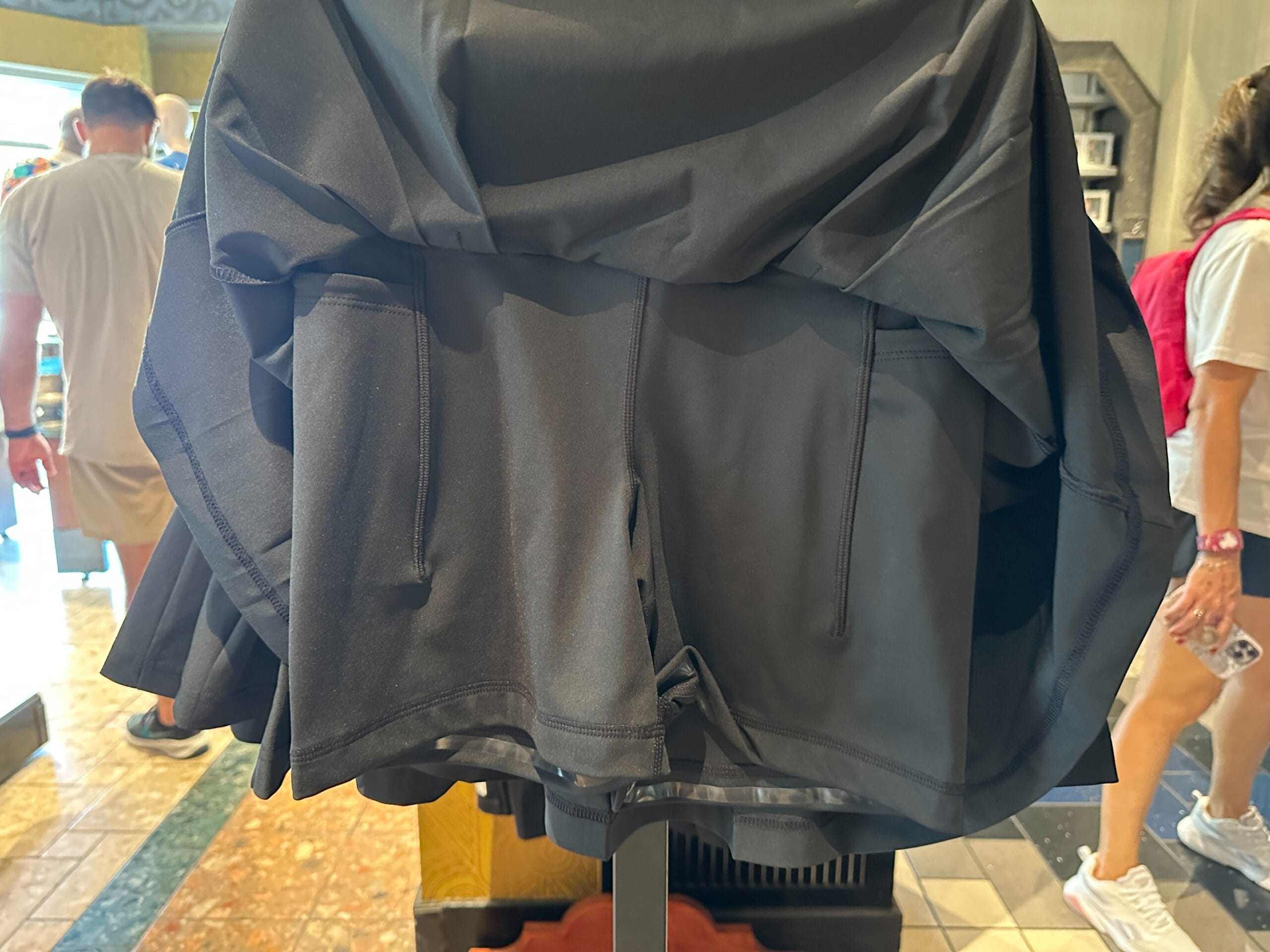 A black hooded jacket from WDW Merch displayed on a rack in a store, with people walking in the background.