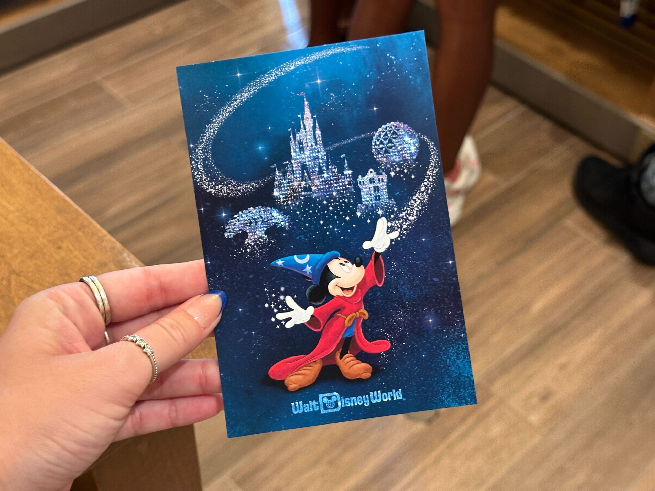 A hand holds WDW Merch—a Walt Disney World postcard featuring Mickey Mouse as a sorcerer and a sparkling castle.