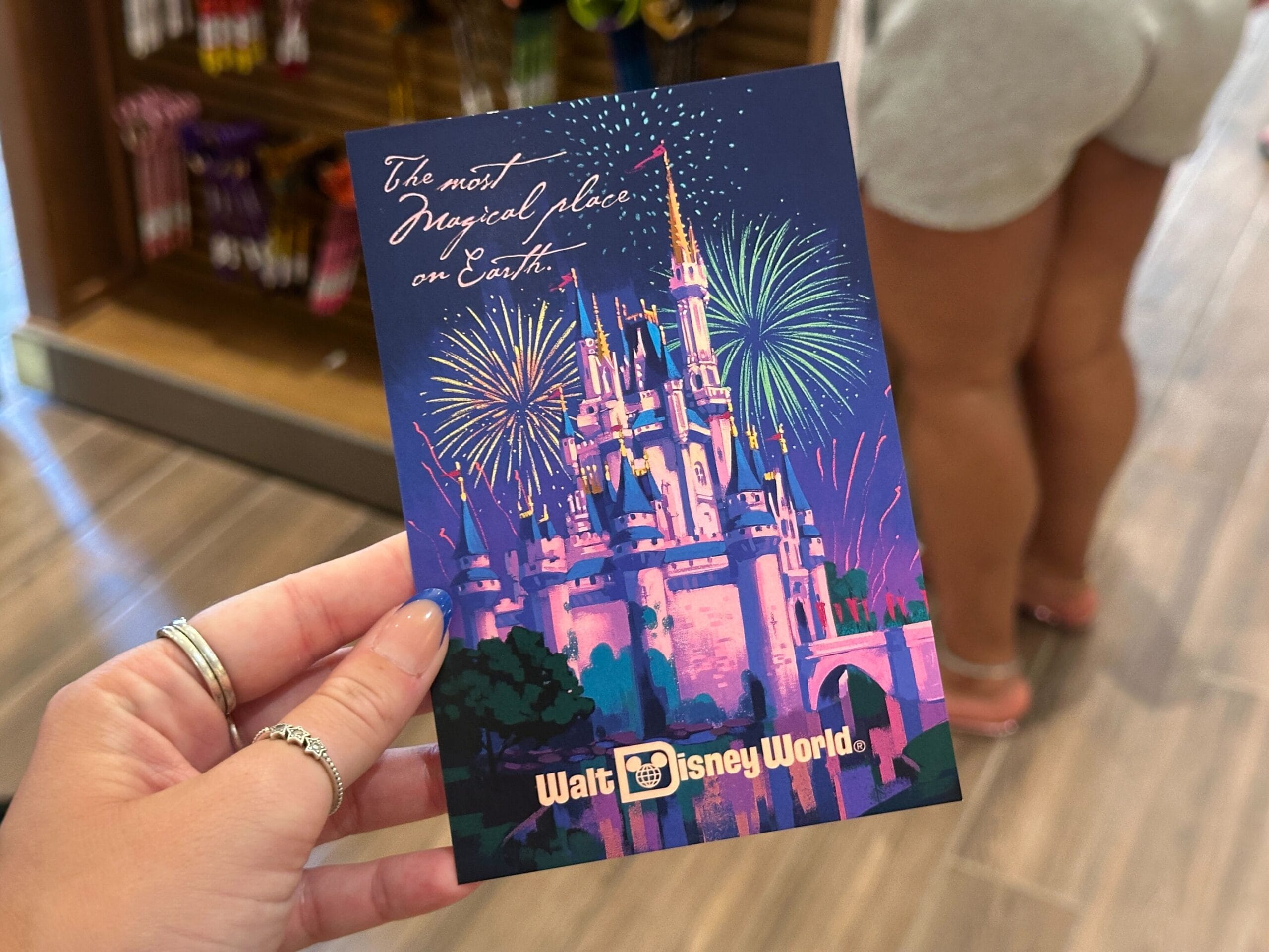 A hand holds WDW merch—a Walt Disney World postcard with a castle, fireworks, and “The most magical place on Earth.”