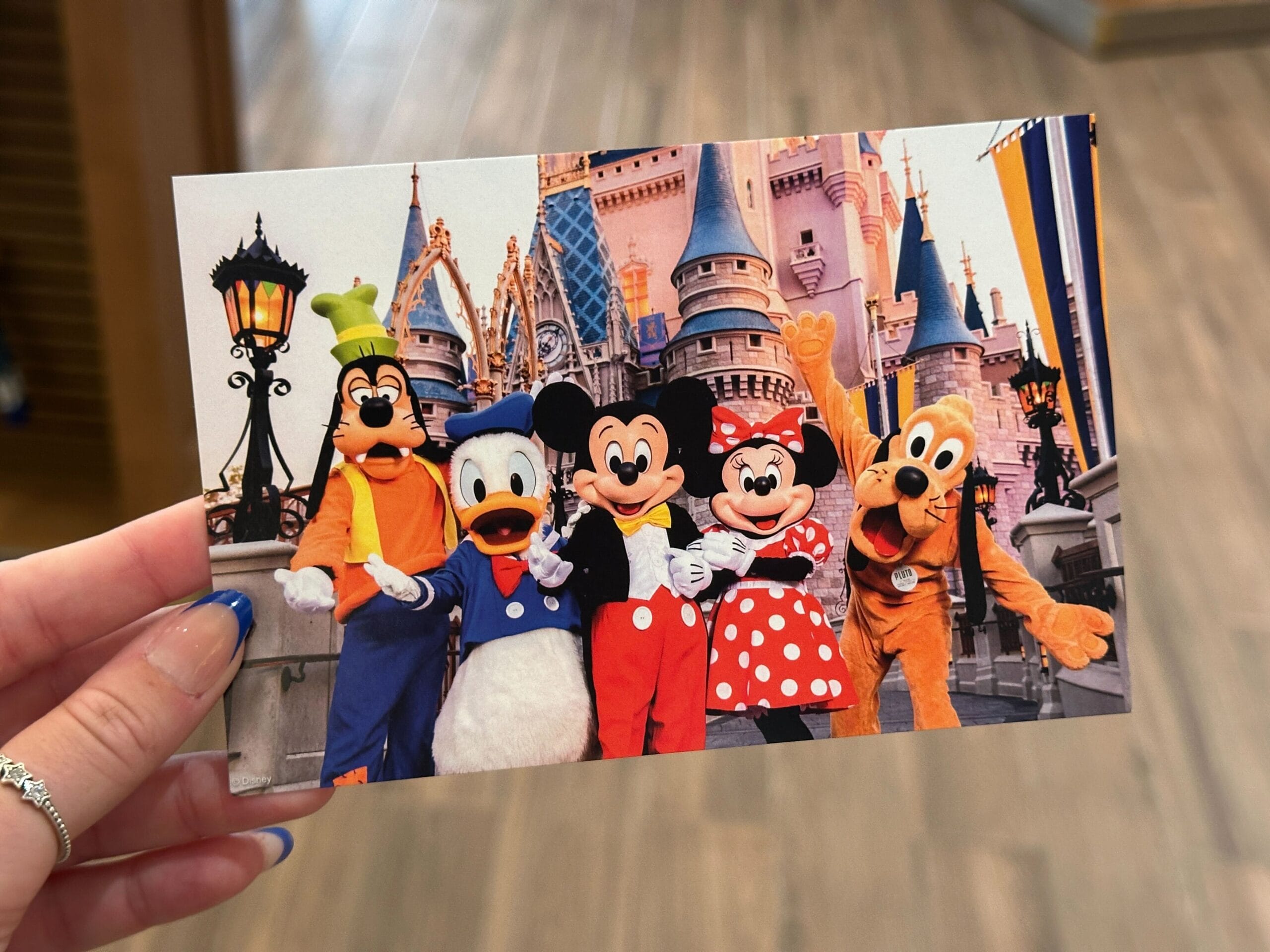 A hand holds a Walt Disney World postcard with Goofy, Donald, Mickey, Minnie, and Pluto in front of the castle.