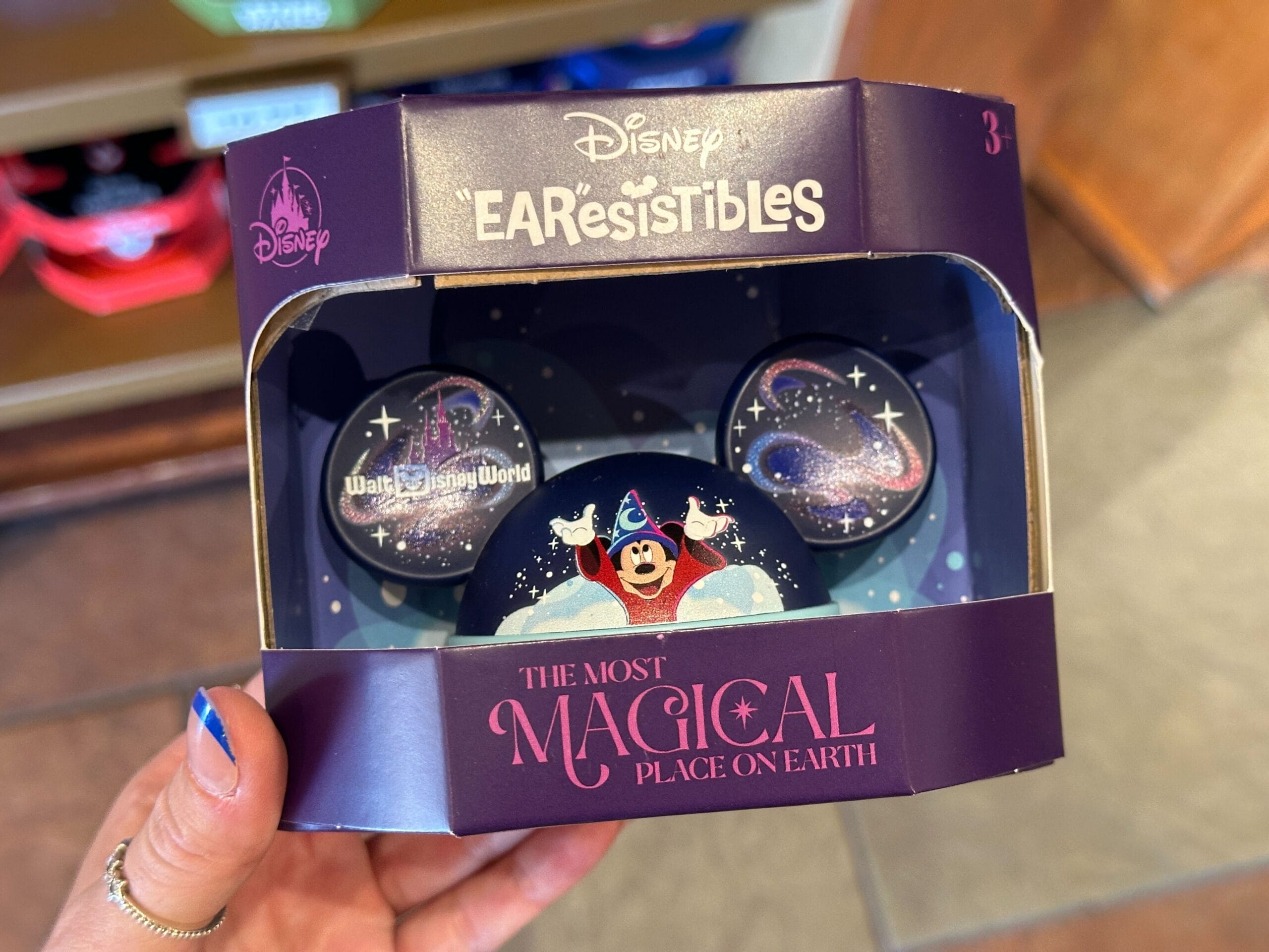 A hand holds WDW Merch: a Disney "EAR-resistibles" ear hat with Sorcerer Mickey and "The Most Magical Place on Earth" text.