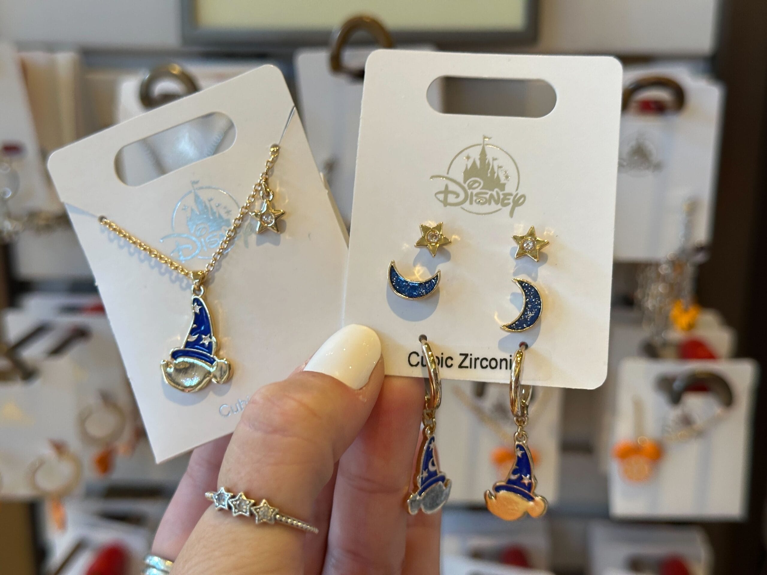 A hand holds Disney jewelry: a blue wizard hat necklace, and matching earrings with stars and crescent moons.