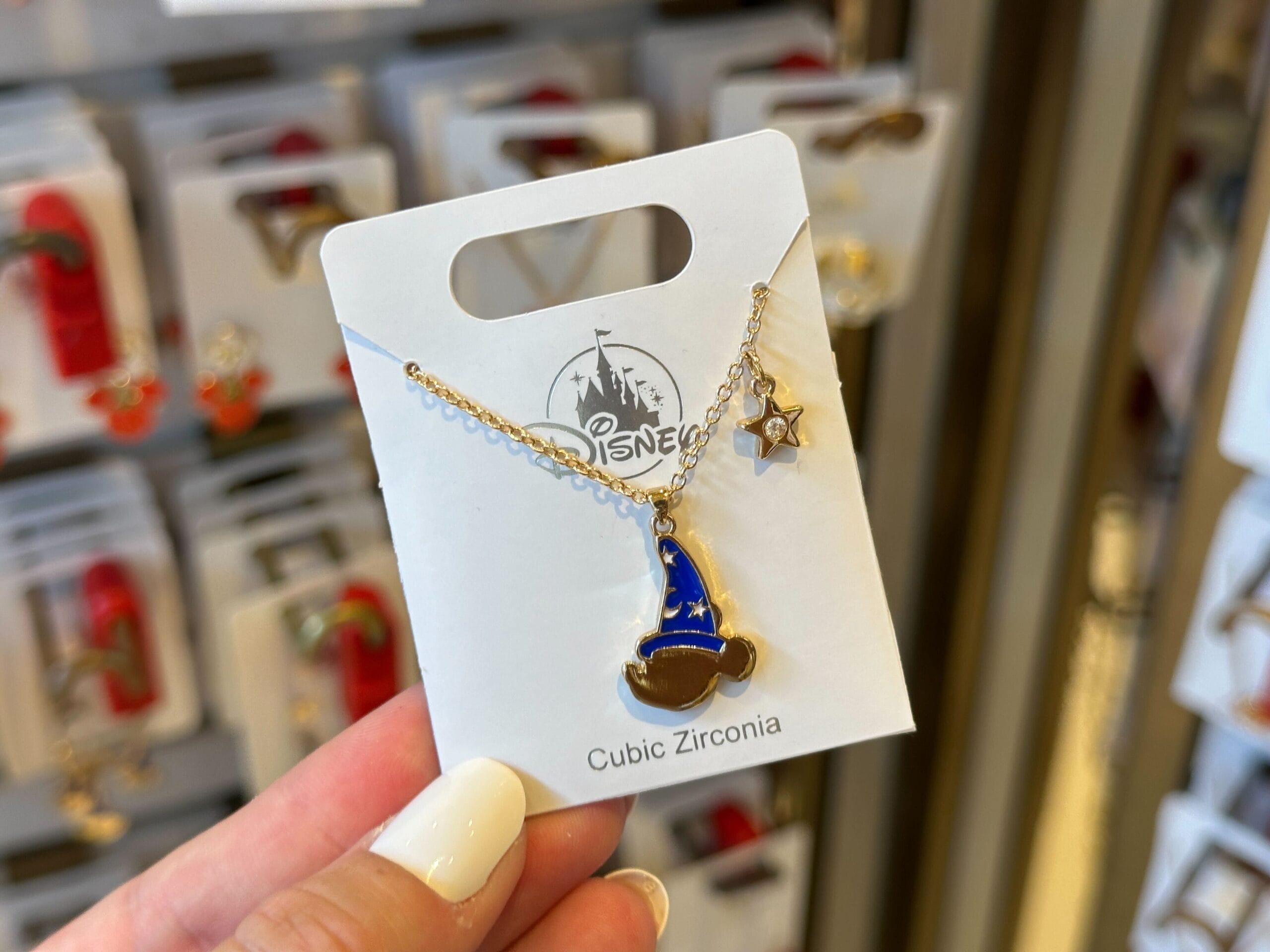 A hand holds a Disney necklace with a blue wizard hat charm and a star charm on a white card.