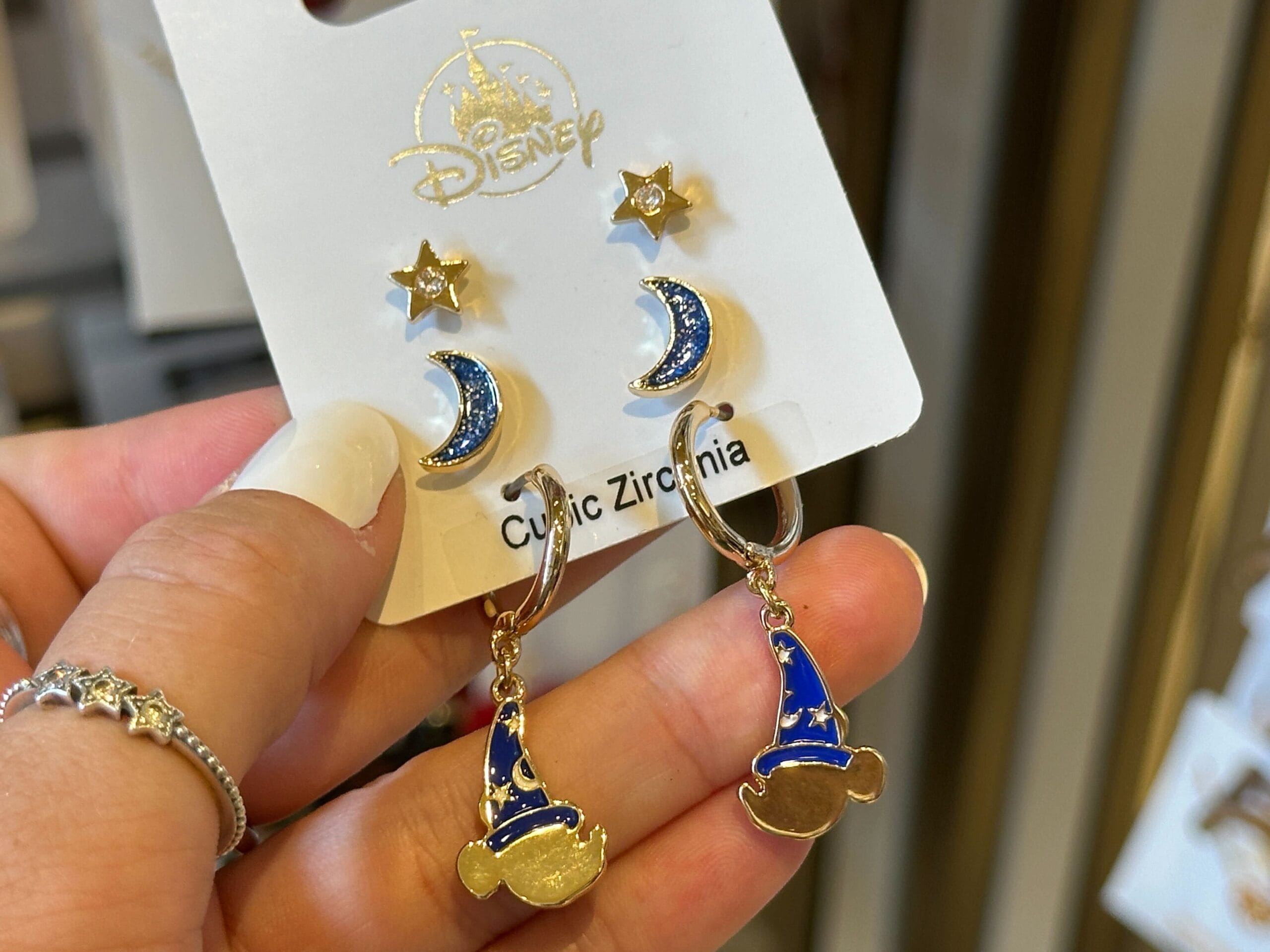 Hand holding Disney earrings: gold stars, blue crescent moons, and blue sorcerer hats with gold Mickey ears.