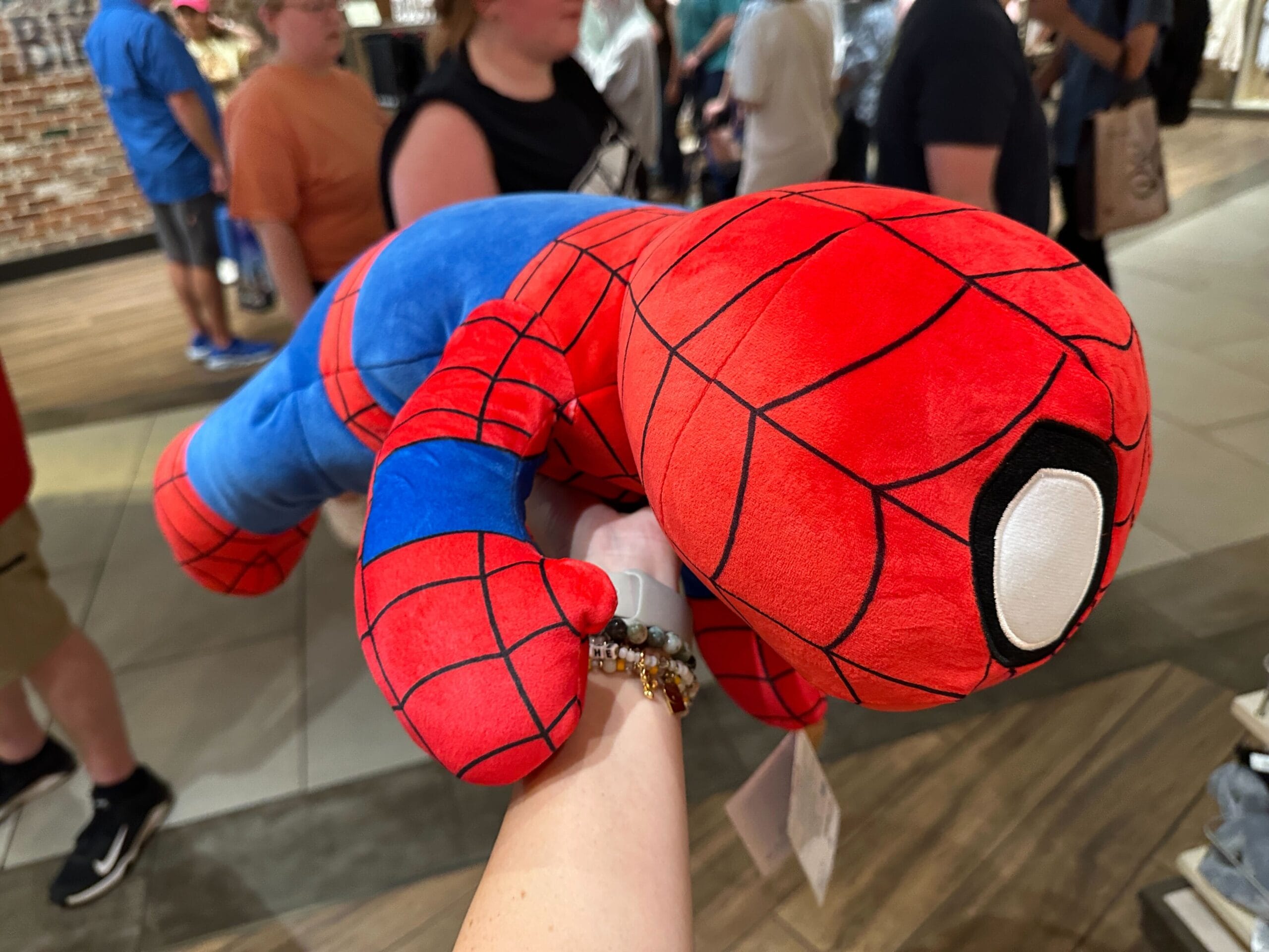A person holds a large, plush Spider-Man toy in a busy indoor shopping area.