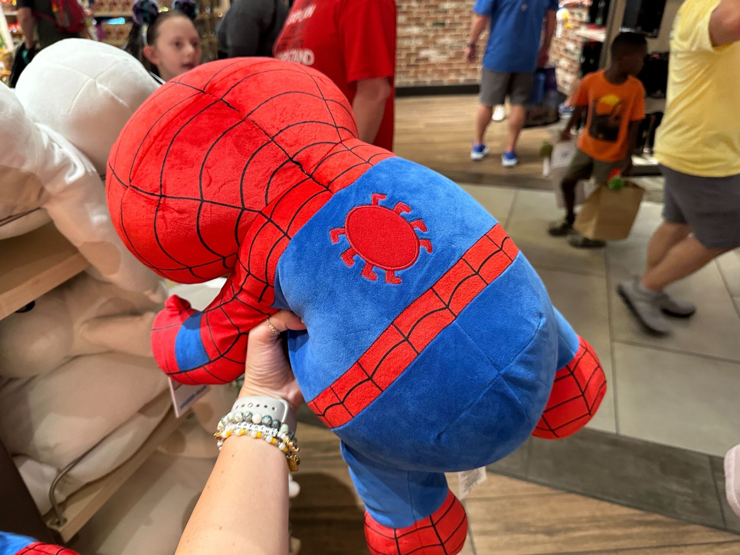 Person holding a plush Spiderman toy in a store with people and shelves in the background.