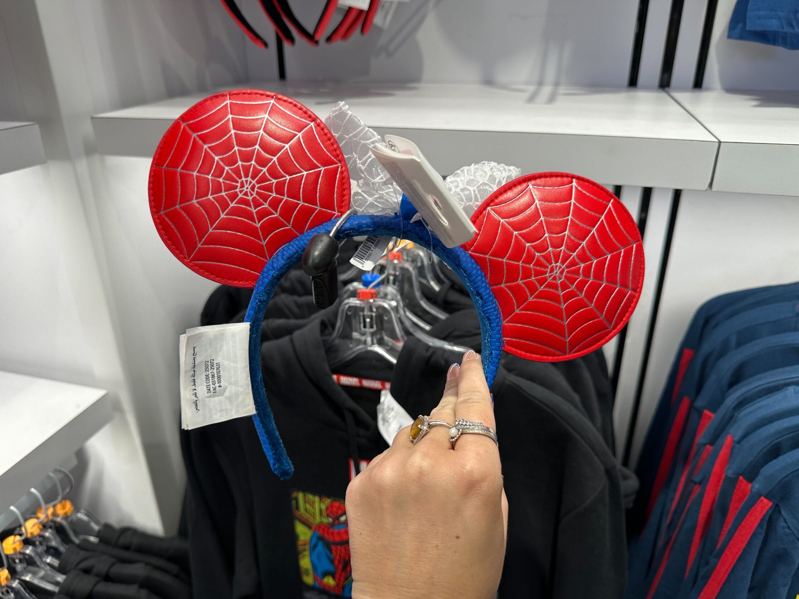 A hand holds a red Mickey Mouse ear headband with a Spider-Man web design among Marvel merchandise in a clothing store.