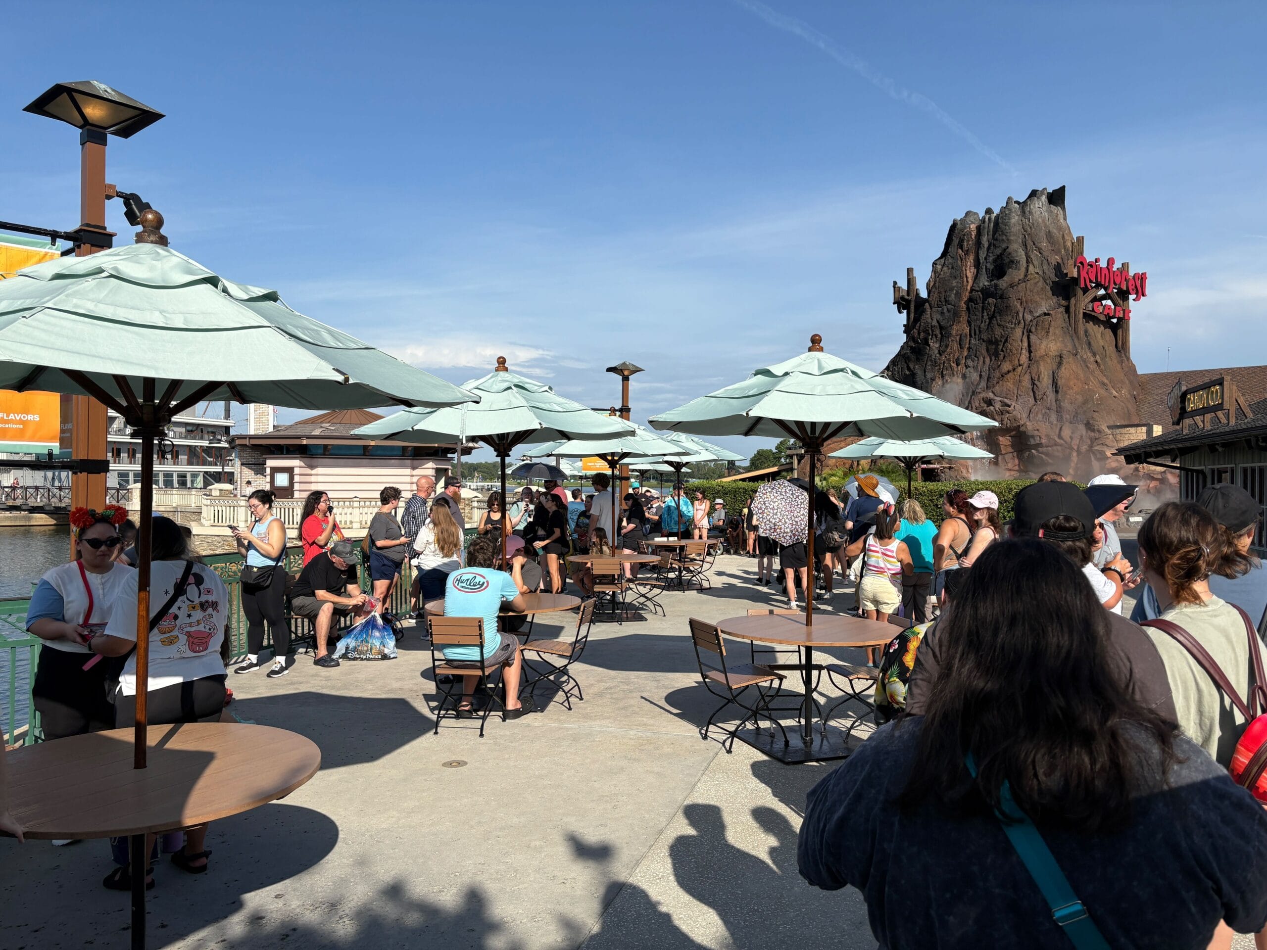 People gathering under umbrellas near outdoor tables on a sunny day, with a volcano-shaped building in the background.