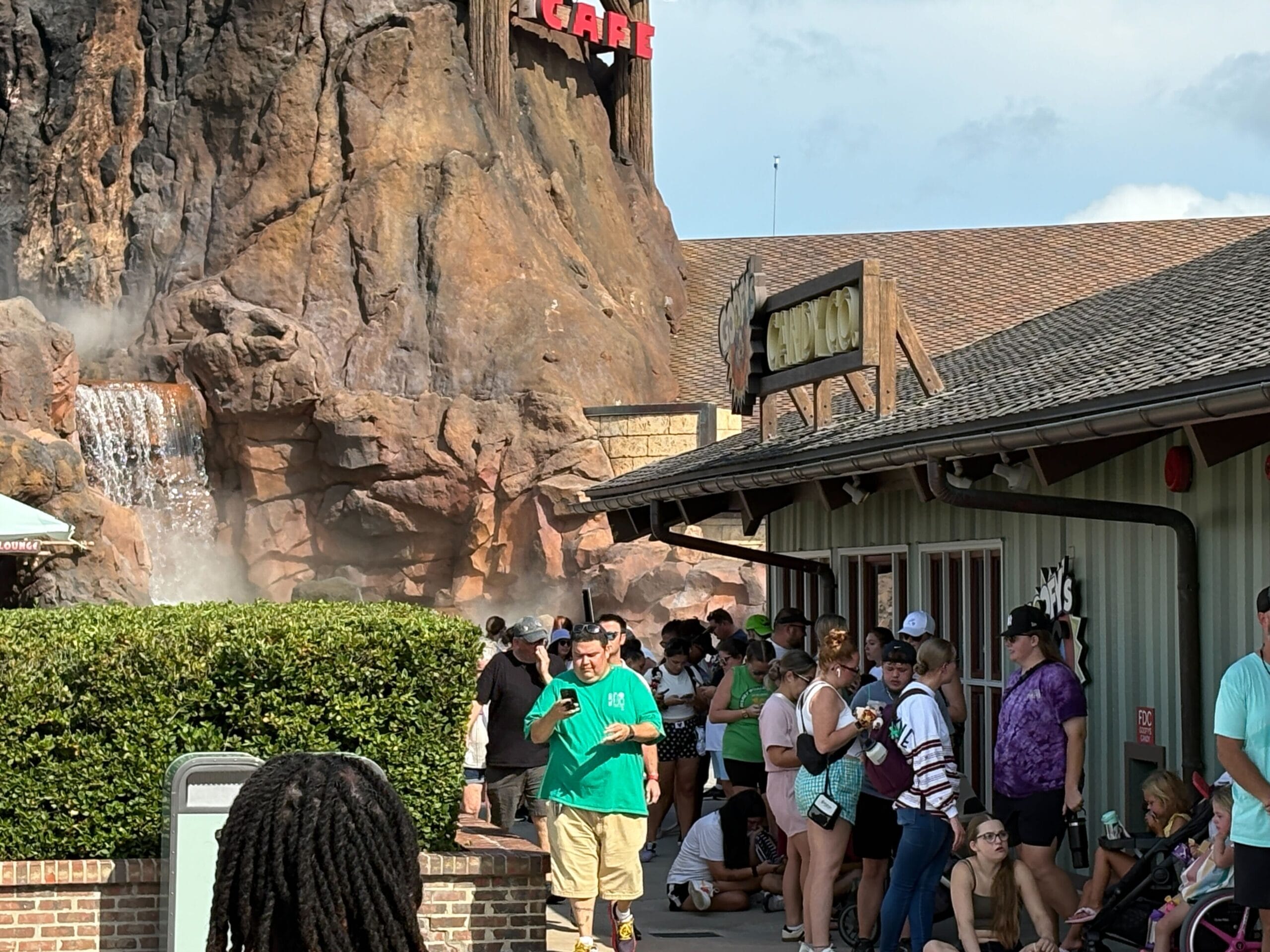 A group of people wait in line outside a building near artificial rocks and a waterfall on a sunny day.