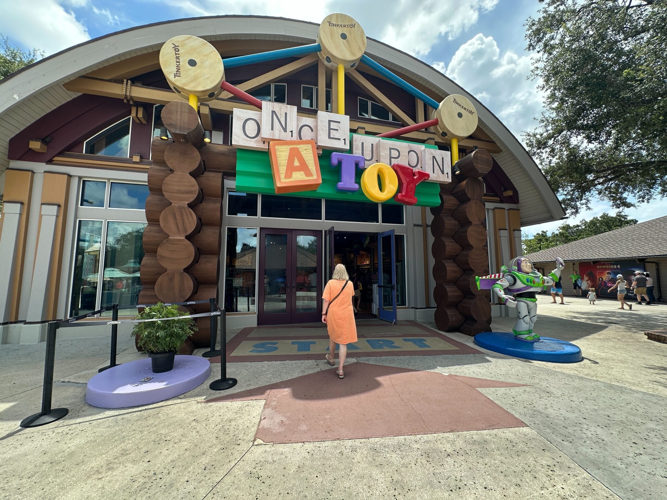 Person in orange dress enters "Once Upon a Toy" store with Buzz Lightyear statue by entrance, under a sunny sky.