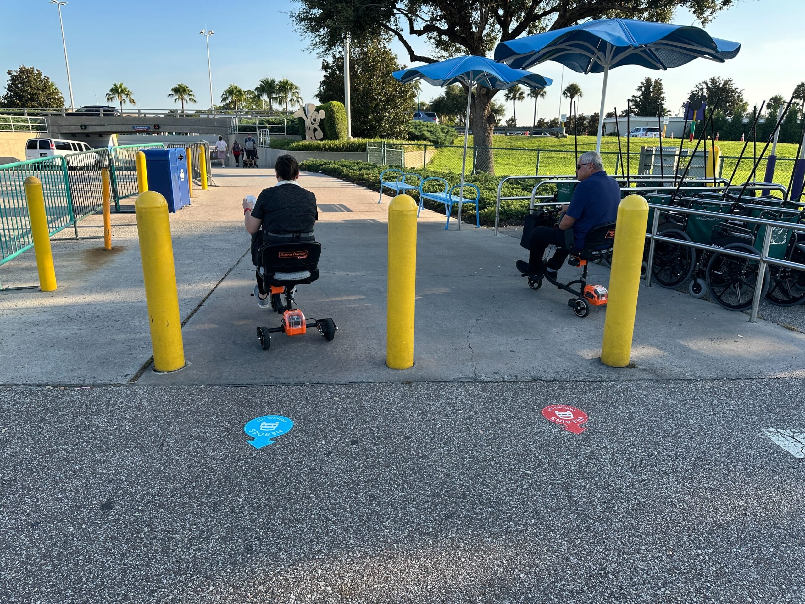 Two people on mobility scooters exit a parking lot, passing yellow poles and wheelchair symbols on the ground.