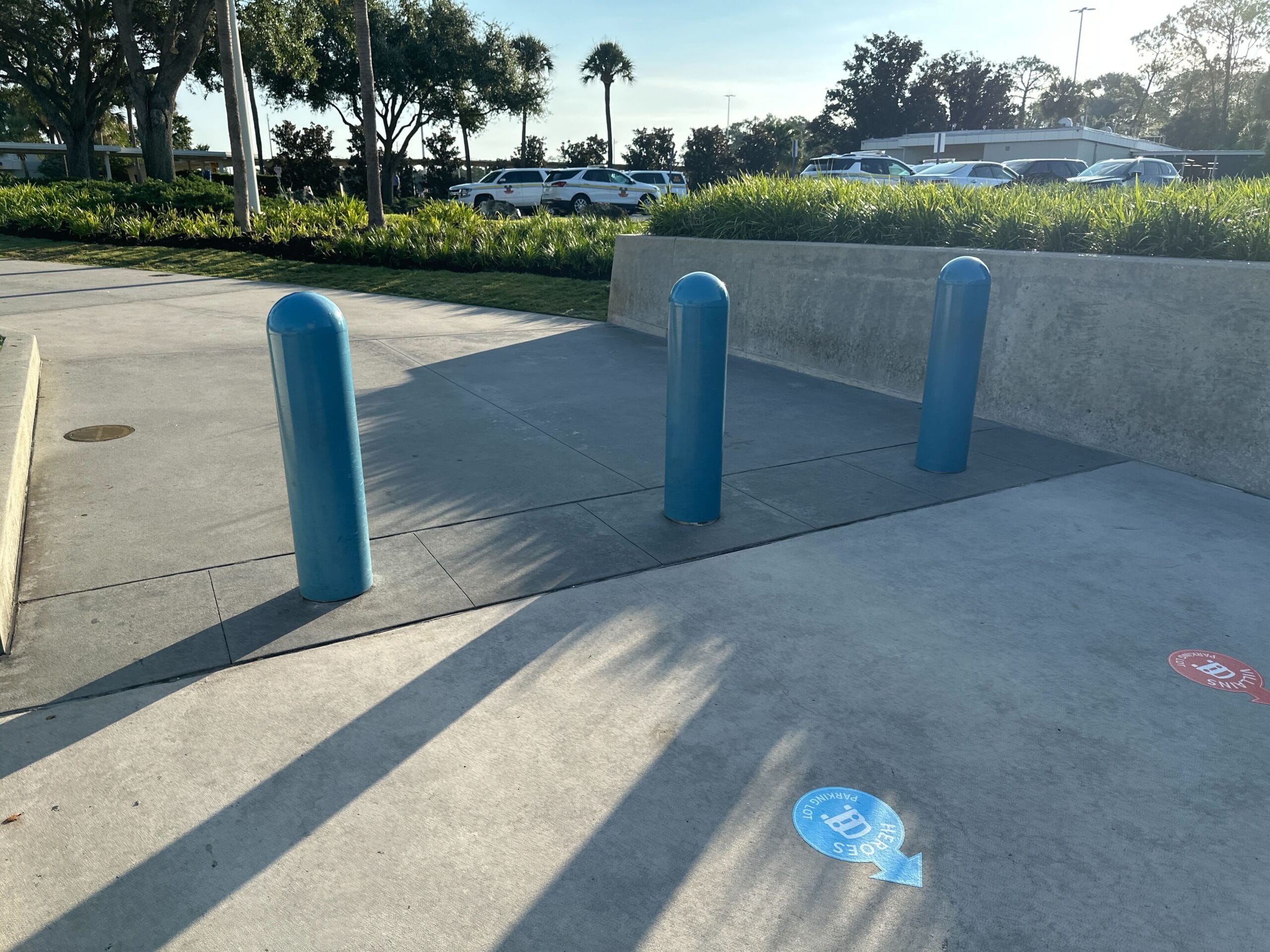 Three blue bollards block a sidewalk entrance surrounded by grass, with cars parked in the background.