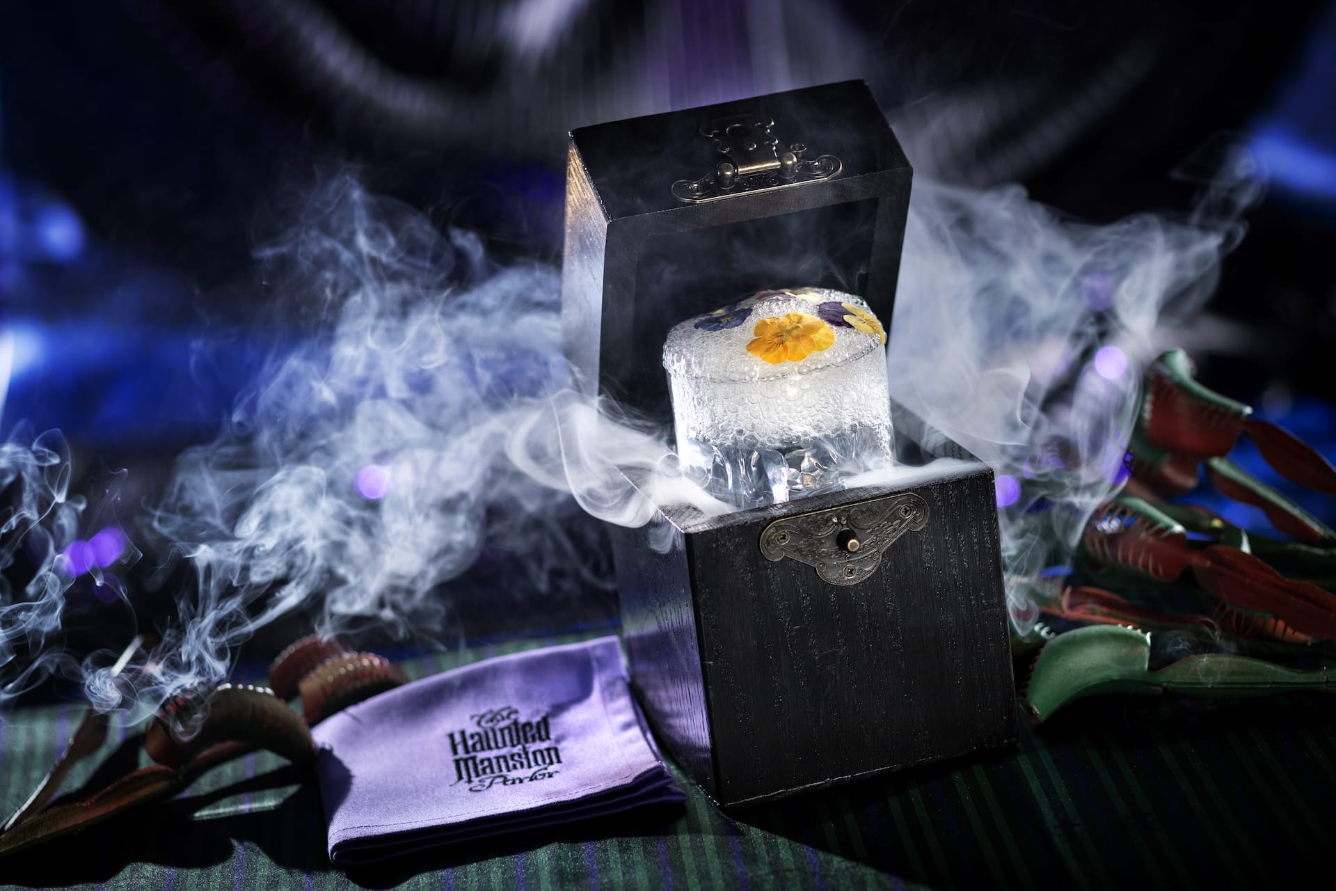 A smoky Disney Destiny cocktail in a black box sits by a purple Haunted Mansion Parlor napkin on a dramatic background.