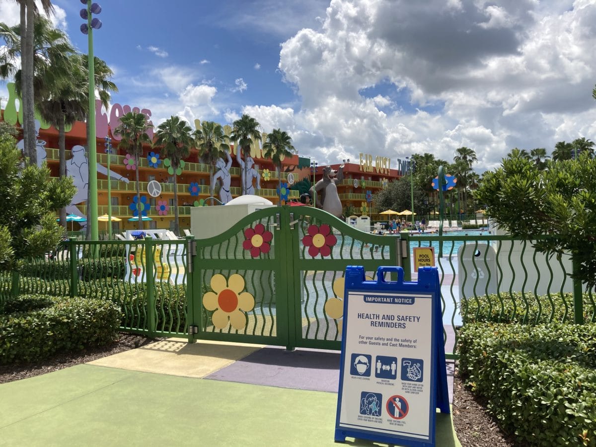 Green gate with flowers by the Hippy Dippy Pool, safety sign in front, palm trees and Disney's Pop Century Resort behind.