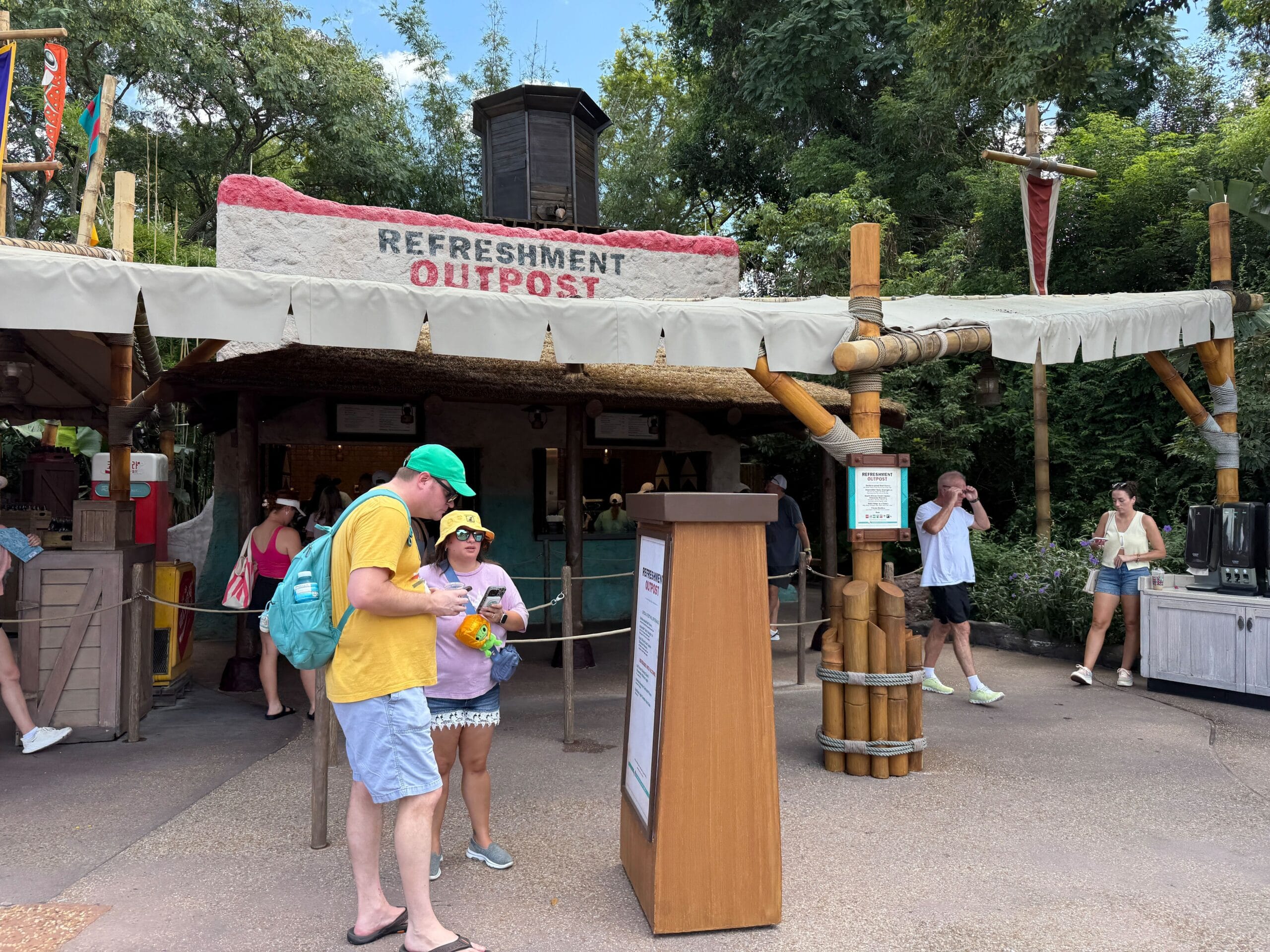 People stand and walk near a "Refreshment Outpost" kiosk at the Food & Wine outdoor venue, surrounded by trees.
