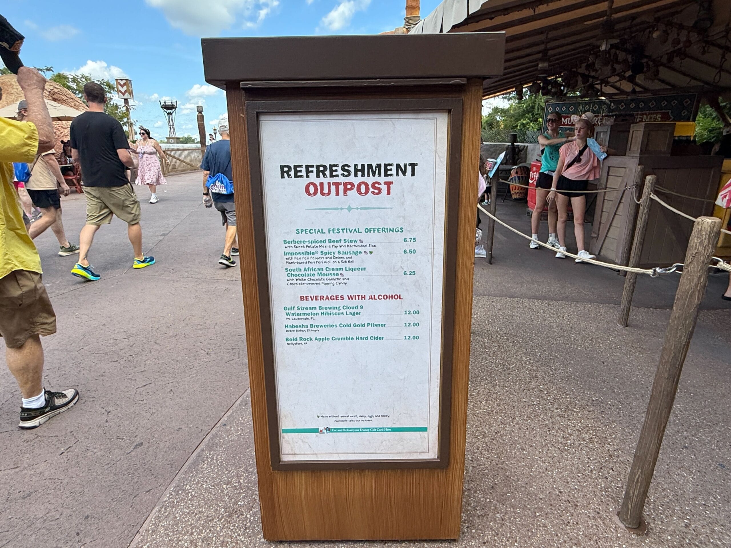 A menu board at the Refreshment Outpost lists Food & Wine options, with people and a building in the background.