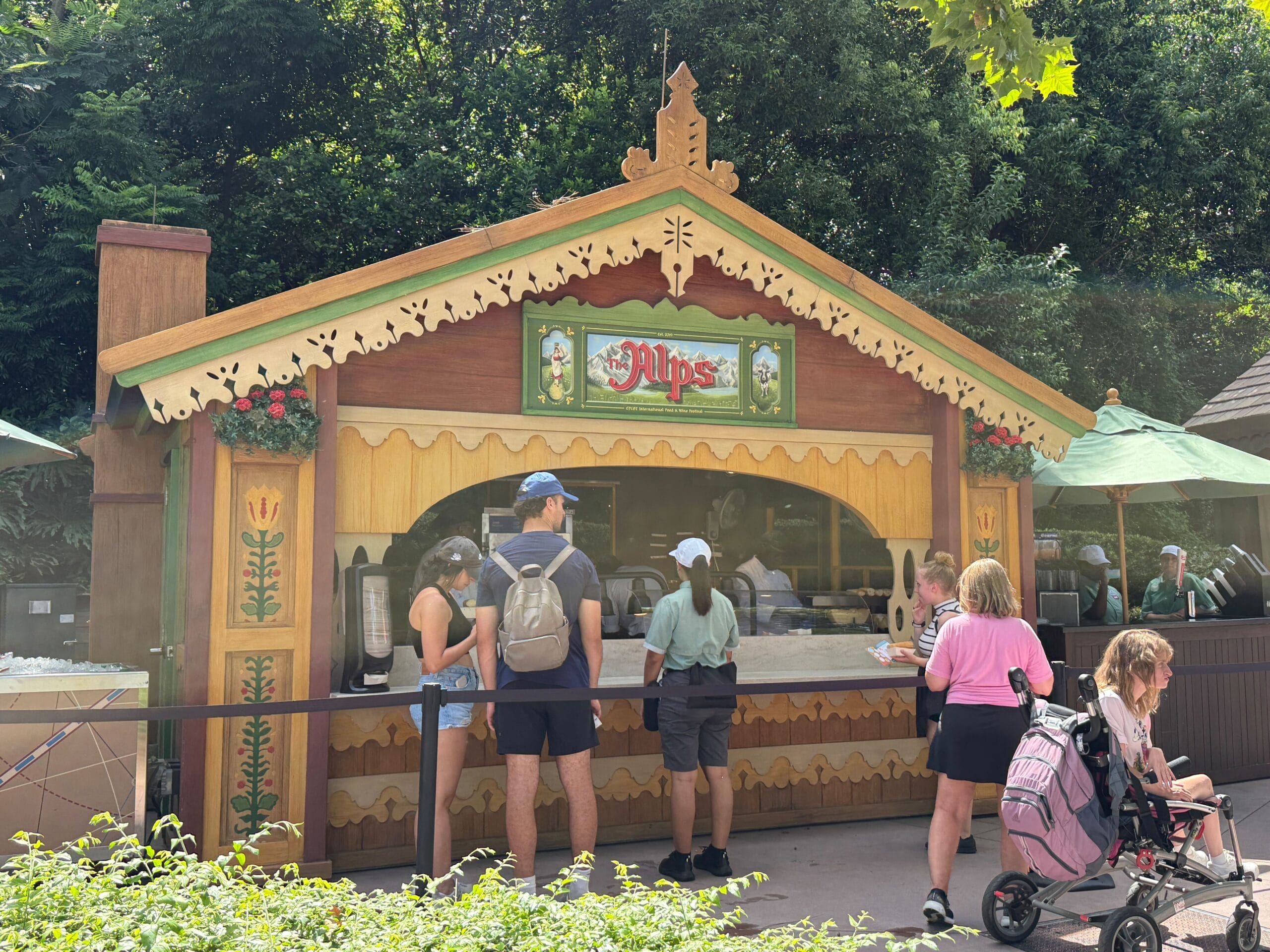 People stand in line at the Alps Food & Wine booth with a decorative wooden facade and green umbrellas outside.