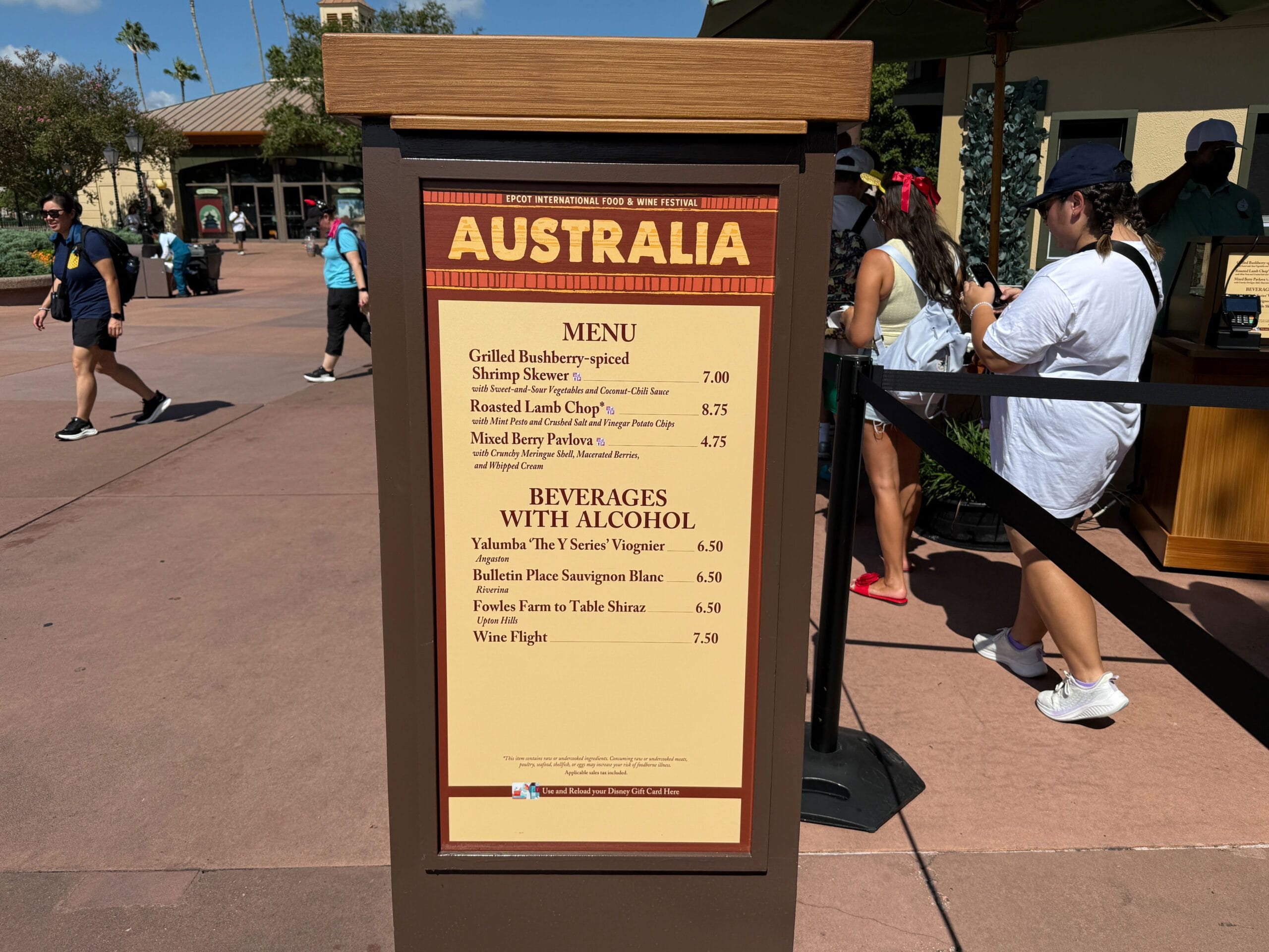 Menu board for Food & Wine booth listing Australian dishes and beverages, with people walking nearby at an outdoor event.