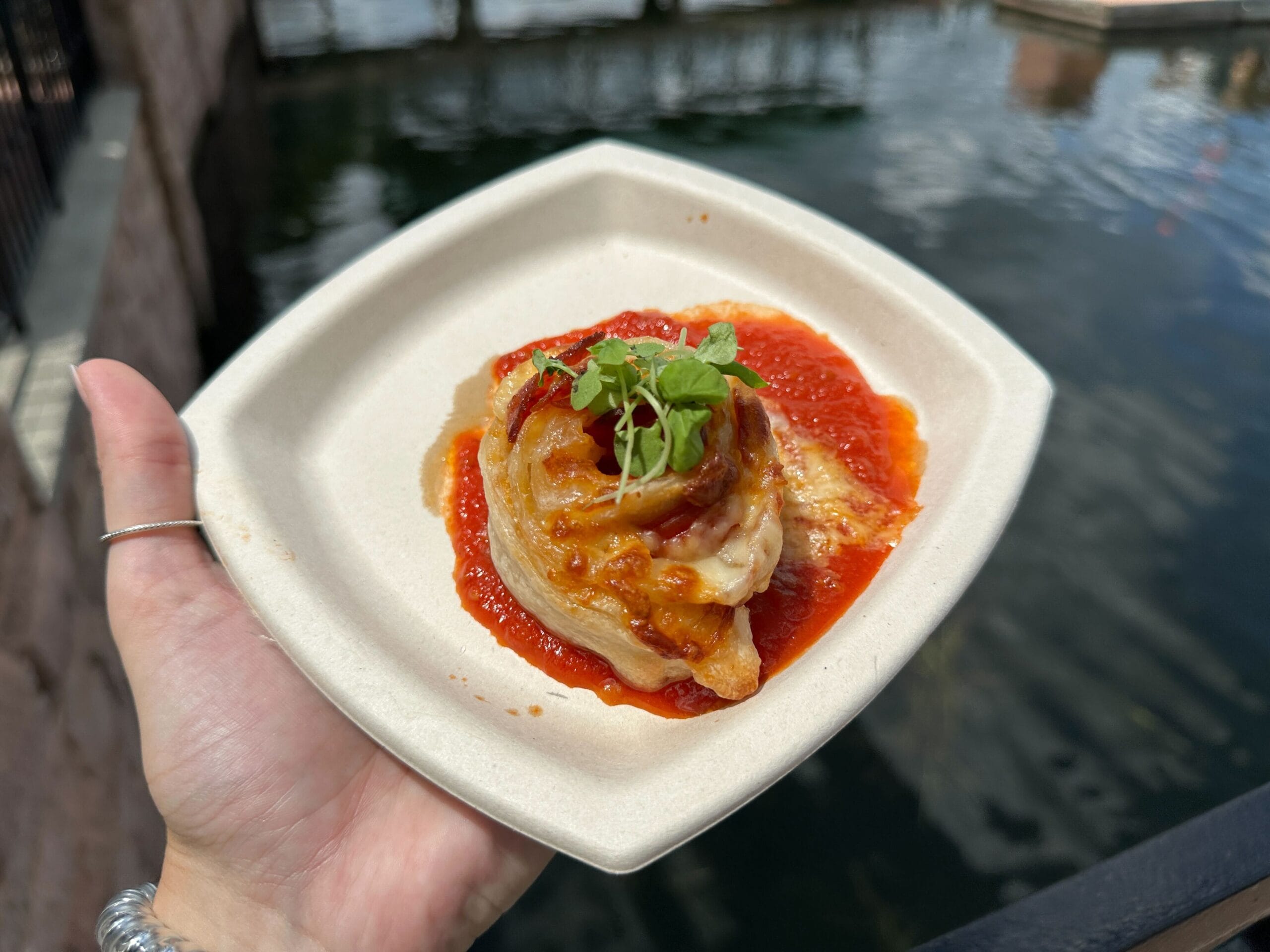 A hand holds a plate with a New Food & Wine stuffed pasta roll topped with sauce and microgreens by the water.