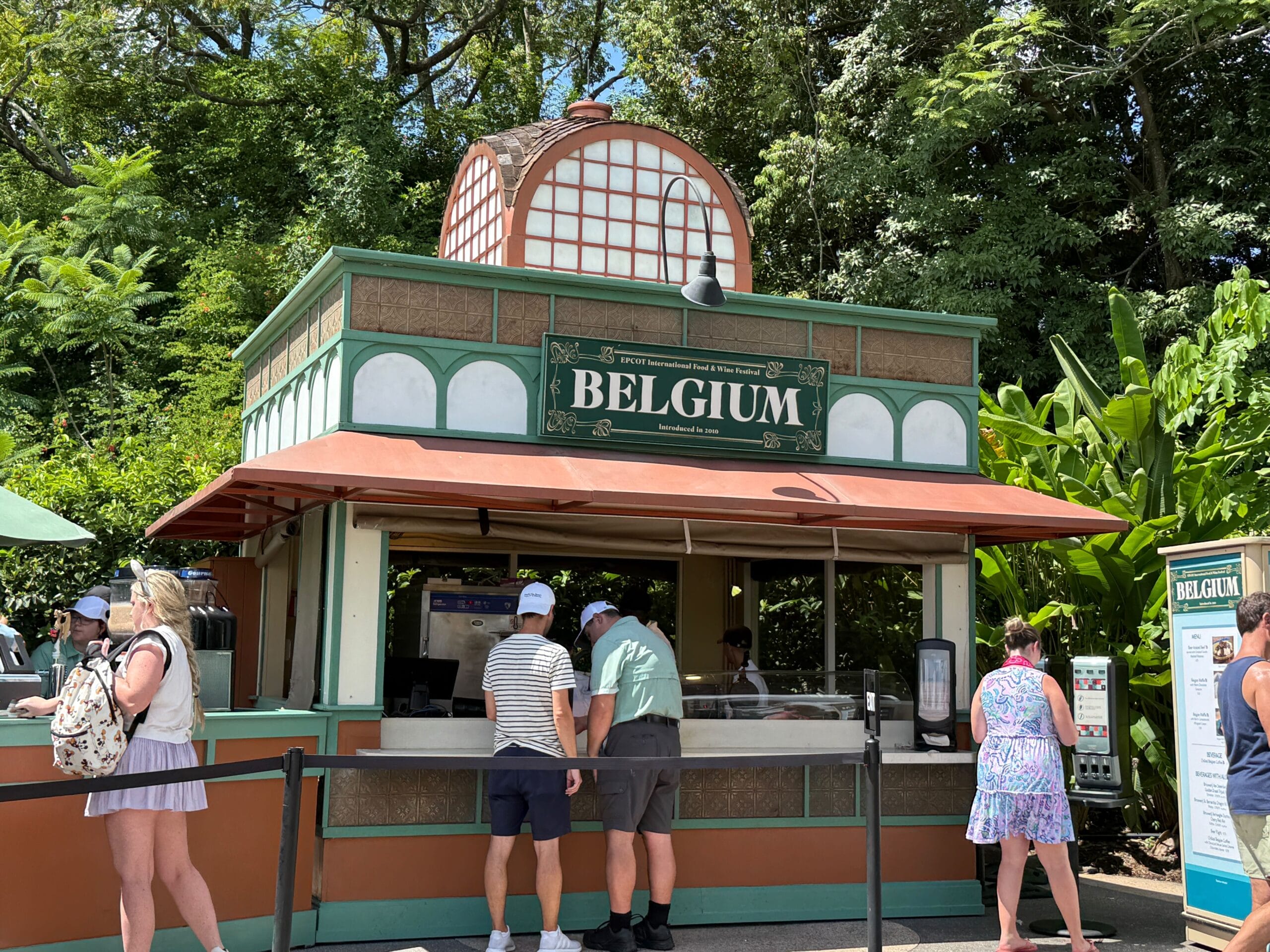 People ordering food & wine at a Belgium-themed outdoor booth surrounded by trees and greenery.